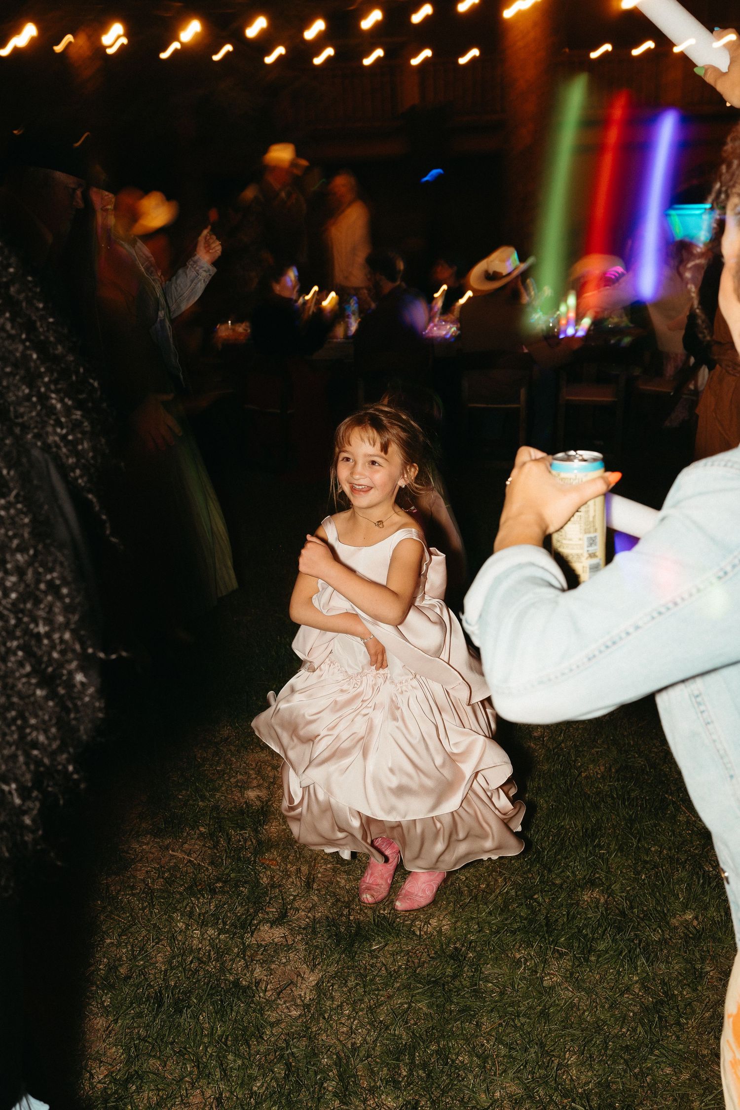 A young girl in a pink dress crouches down on grass at a nighttime outdoor party with colorful lights.