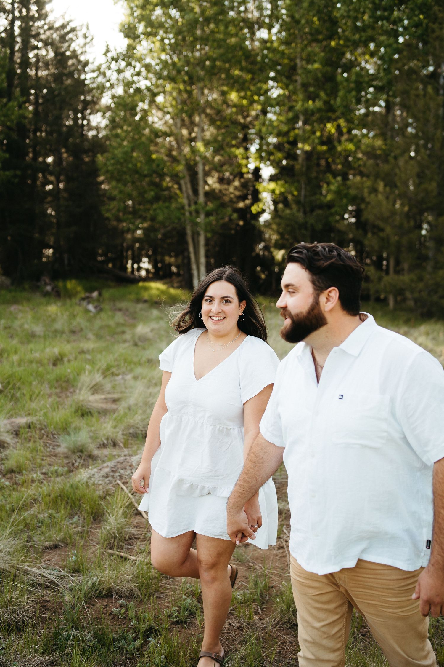Couple wearing white walking together in a grassy forest clearing during golden hour.