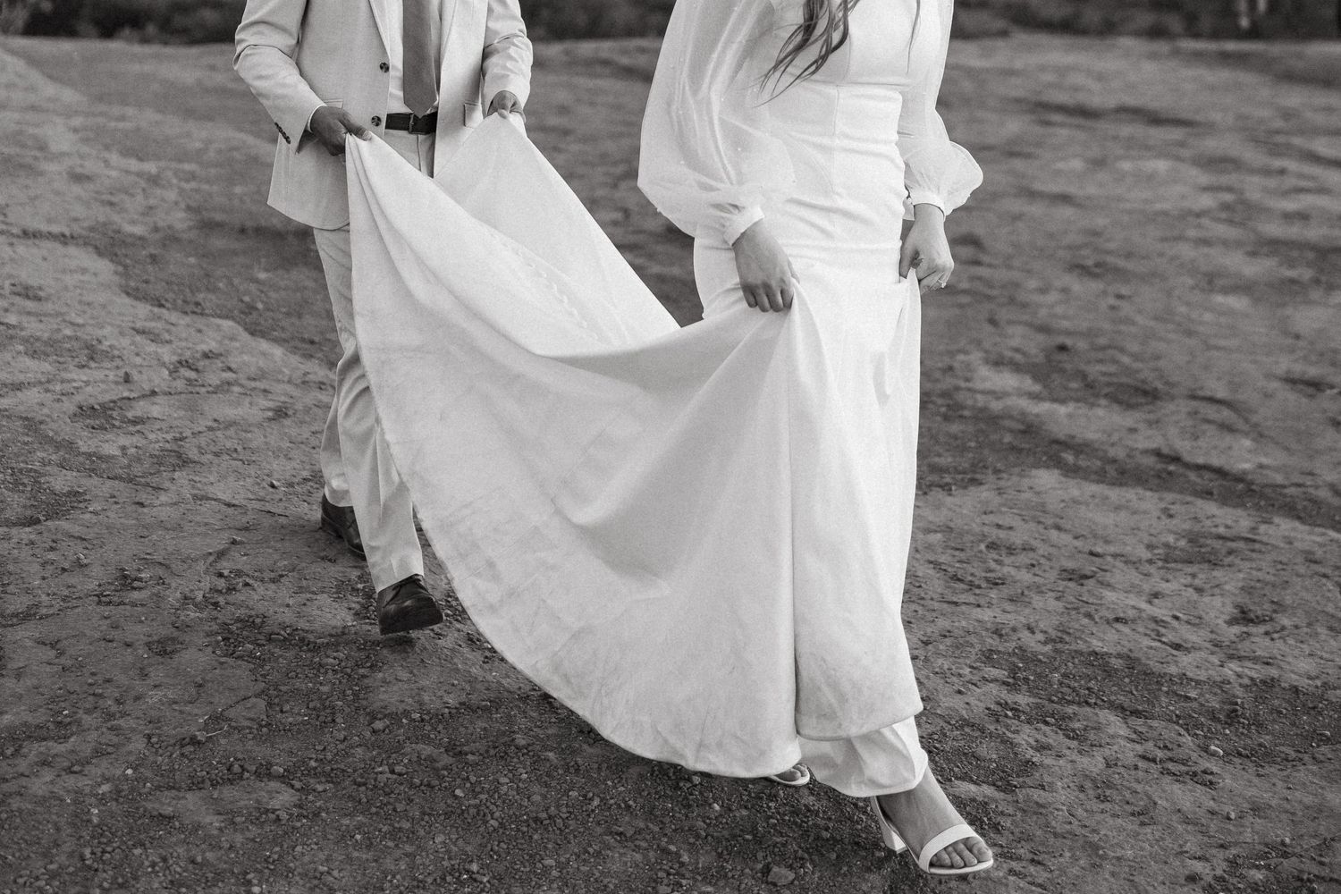 Black and white image of couple holding wedding dress while walking.
