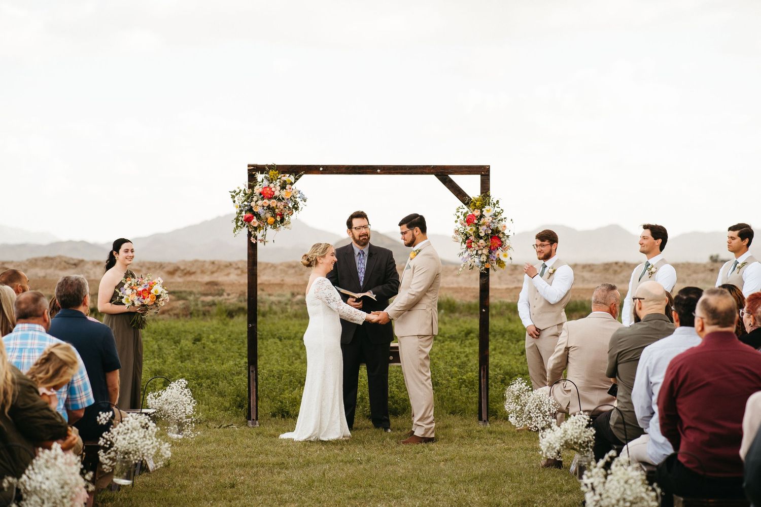 Outdoor wedding ceremony under wooden arch decorated with floral arrangements on hilltop setting.