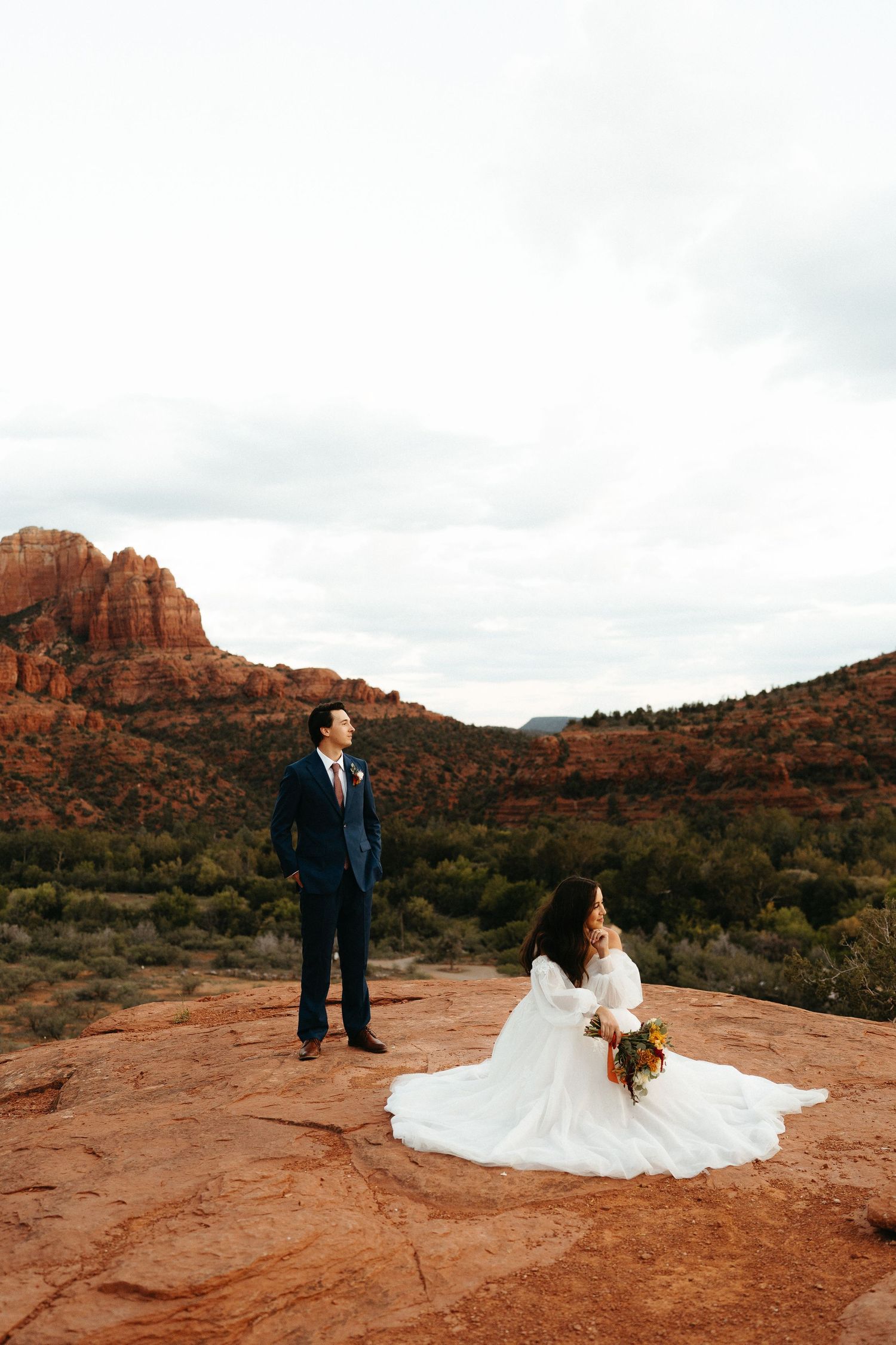 A couple in wedding attire poses on red rock formations in Sedona with dramatic desert landscape in the background.