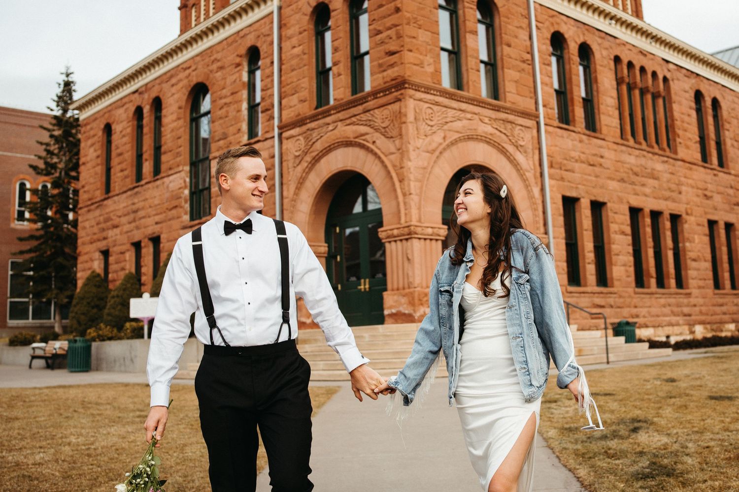 Couple in formal attire walking hand in hand outside historic brick building.