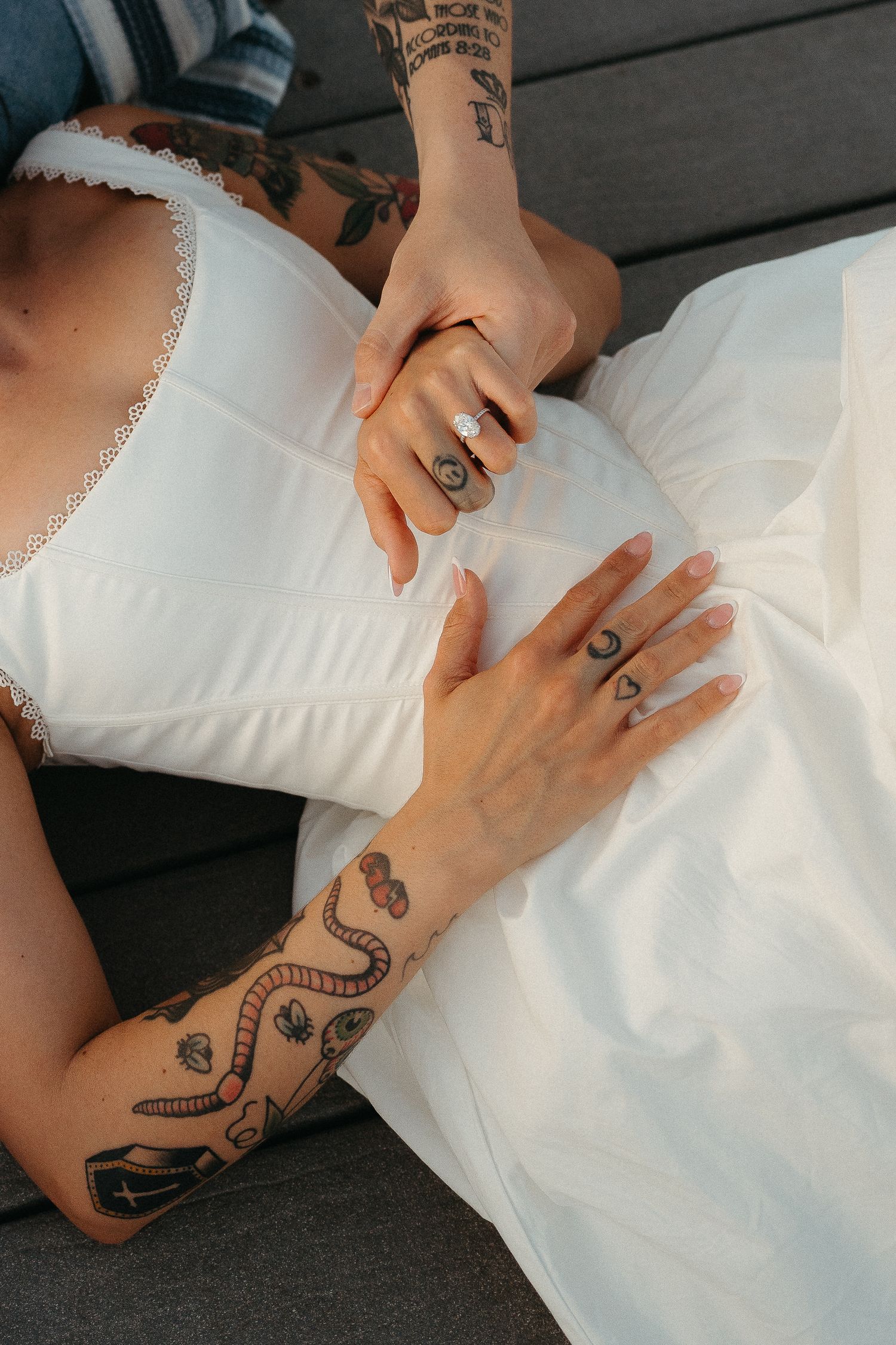 Close-up of tattooed hands resting on white lace dress fabric showing multiple rings and intricate arm tattoo designs.