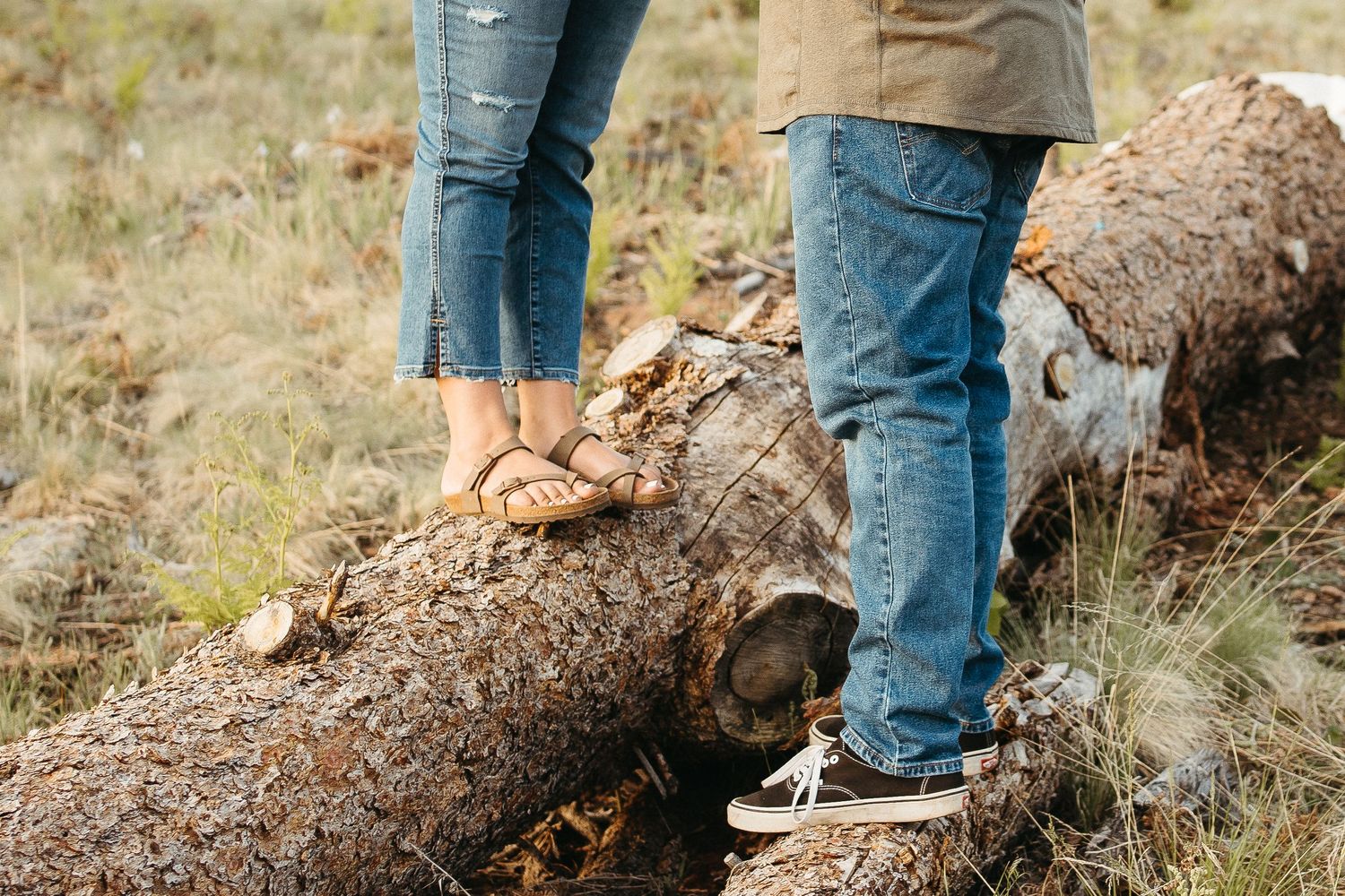 Two pairs of feet in casual wear standing on a fallen tree trunk in nature.
