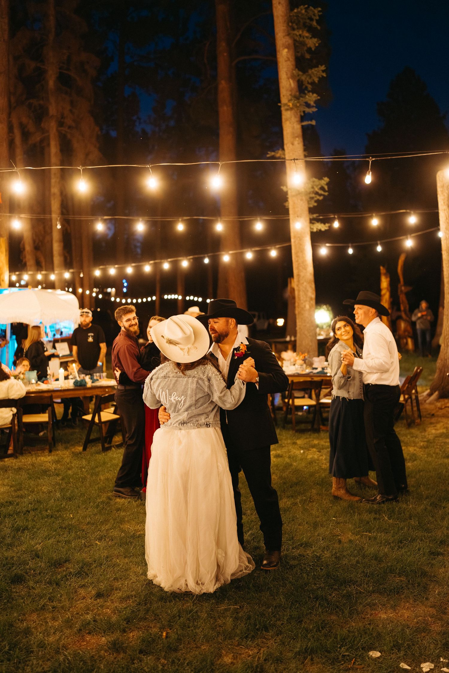 Outdoor evening wedding reception with string lights illuminating couples dancing on a grassy area between trees.