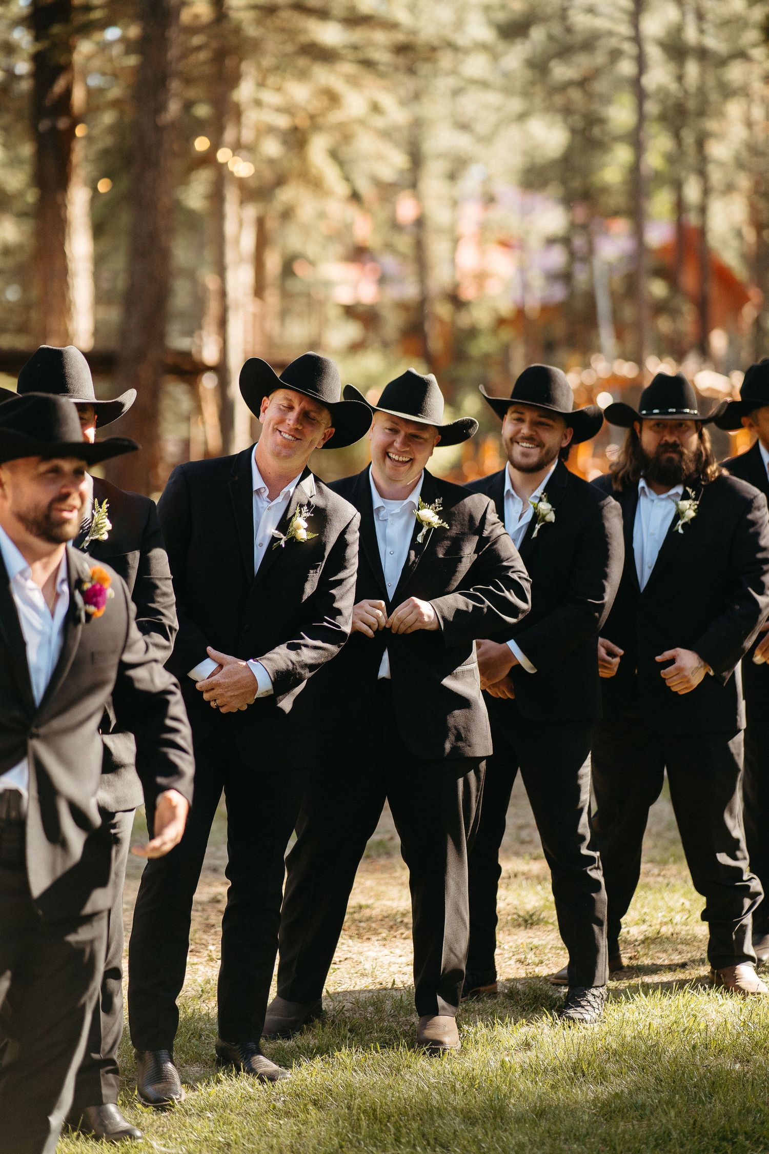 Groomsmen in black suits and cowboy hats line up for a fun photo in a wooded outdoor setting.