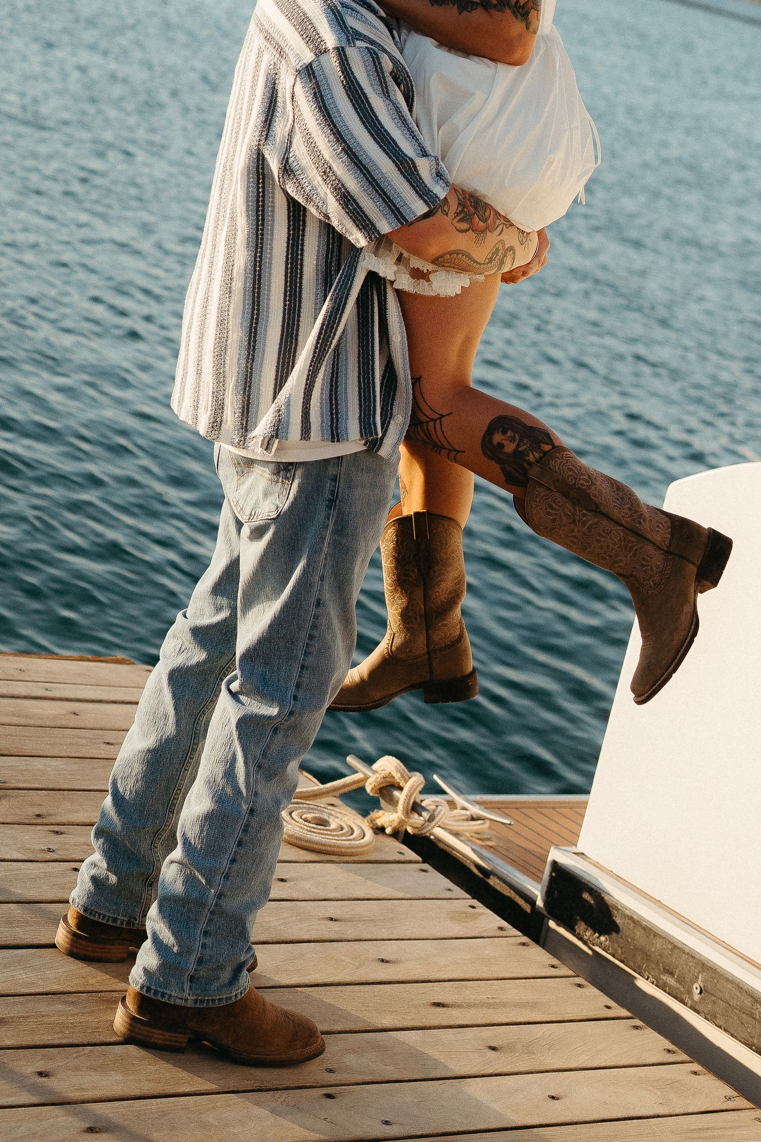 Couple standing on a wooden dock by the water, wearing jeans and boots in a casual romantic embrace.