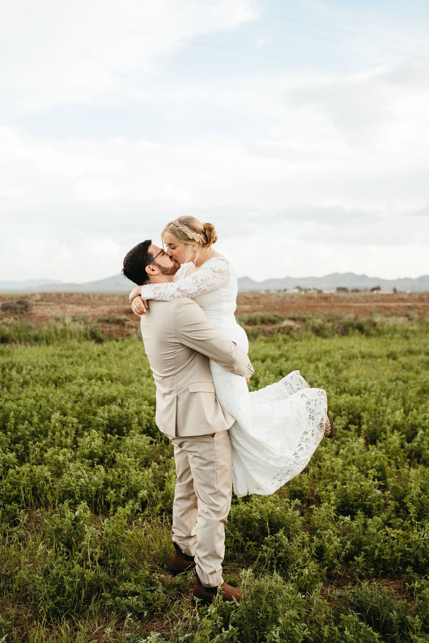Romantic couple embrace in field with mountains in background during sunset wedding photo.