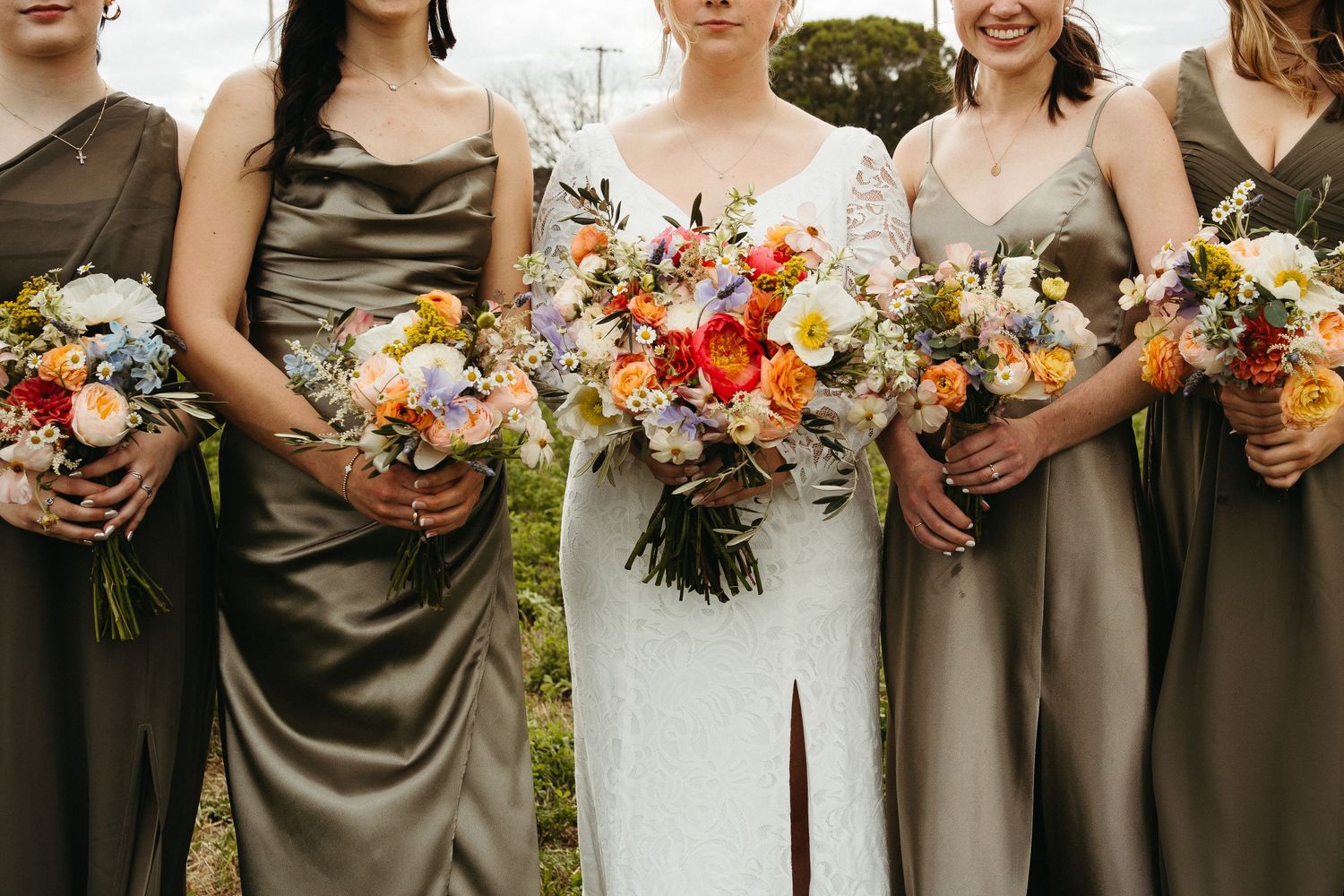 Wedding party displays colorful bouquets featuring orange and white flowers against olive dresses.