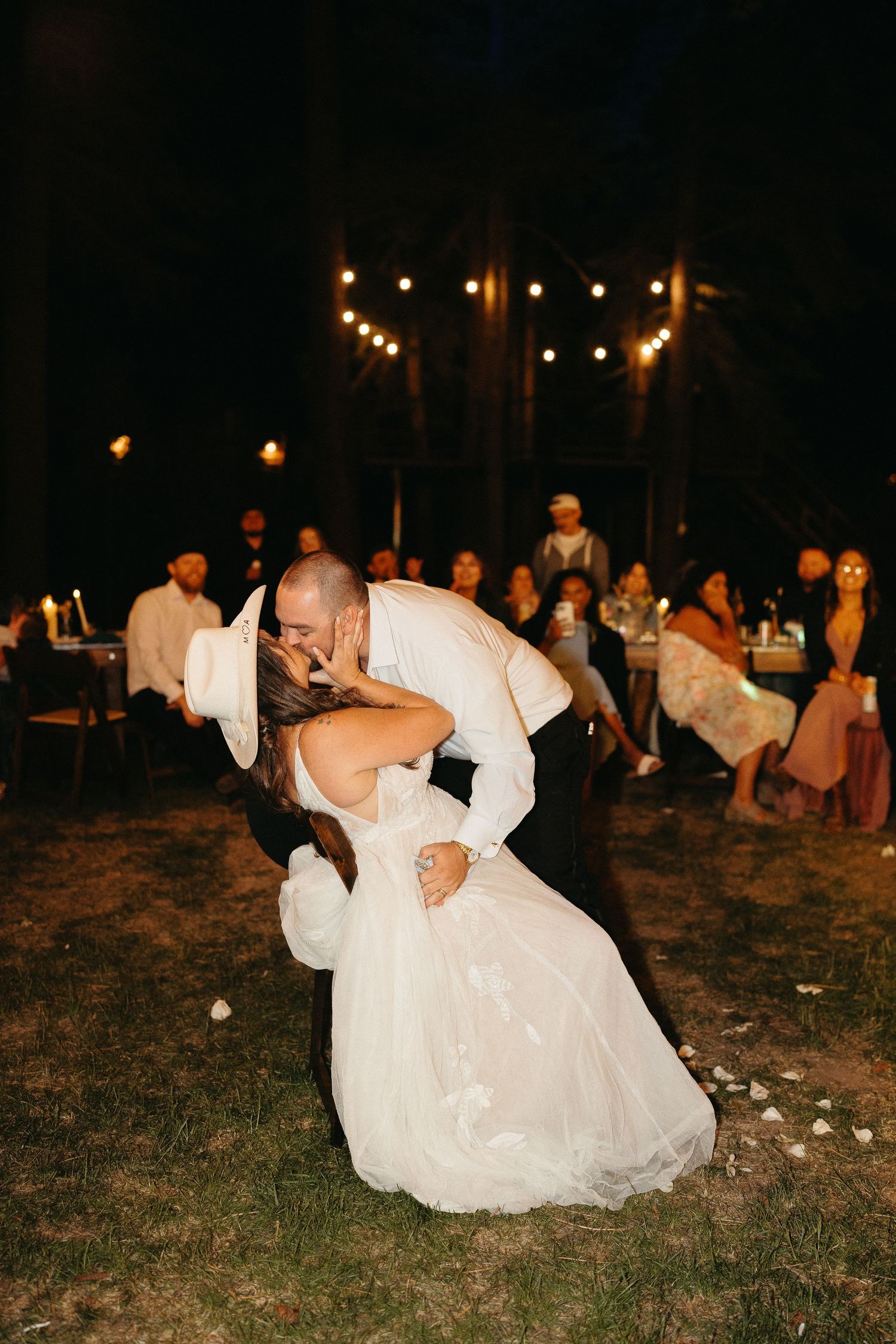 A couple in wedding attire performs a romantic dip dance move on grass at night under string lights.