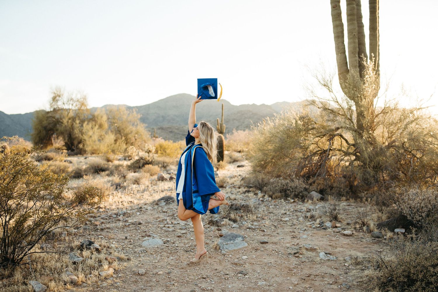 Graduate in blue cap and gown throws tassel in desert landscape with saguaro cactus.
