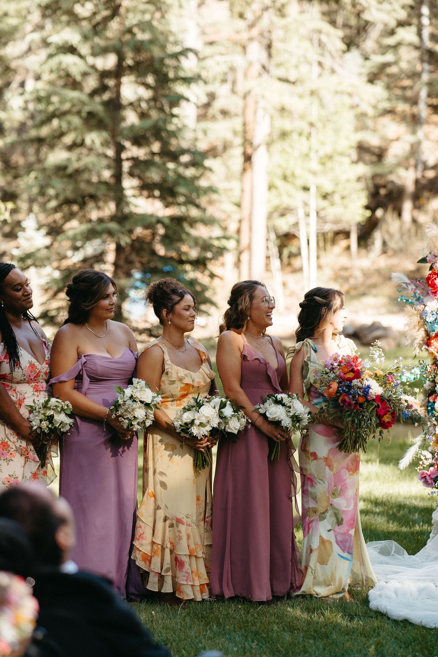 Bridesmaids wearing purple and floral dresses stand in line holding colorful bouquets during an outdoor forest wedding ceremony.