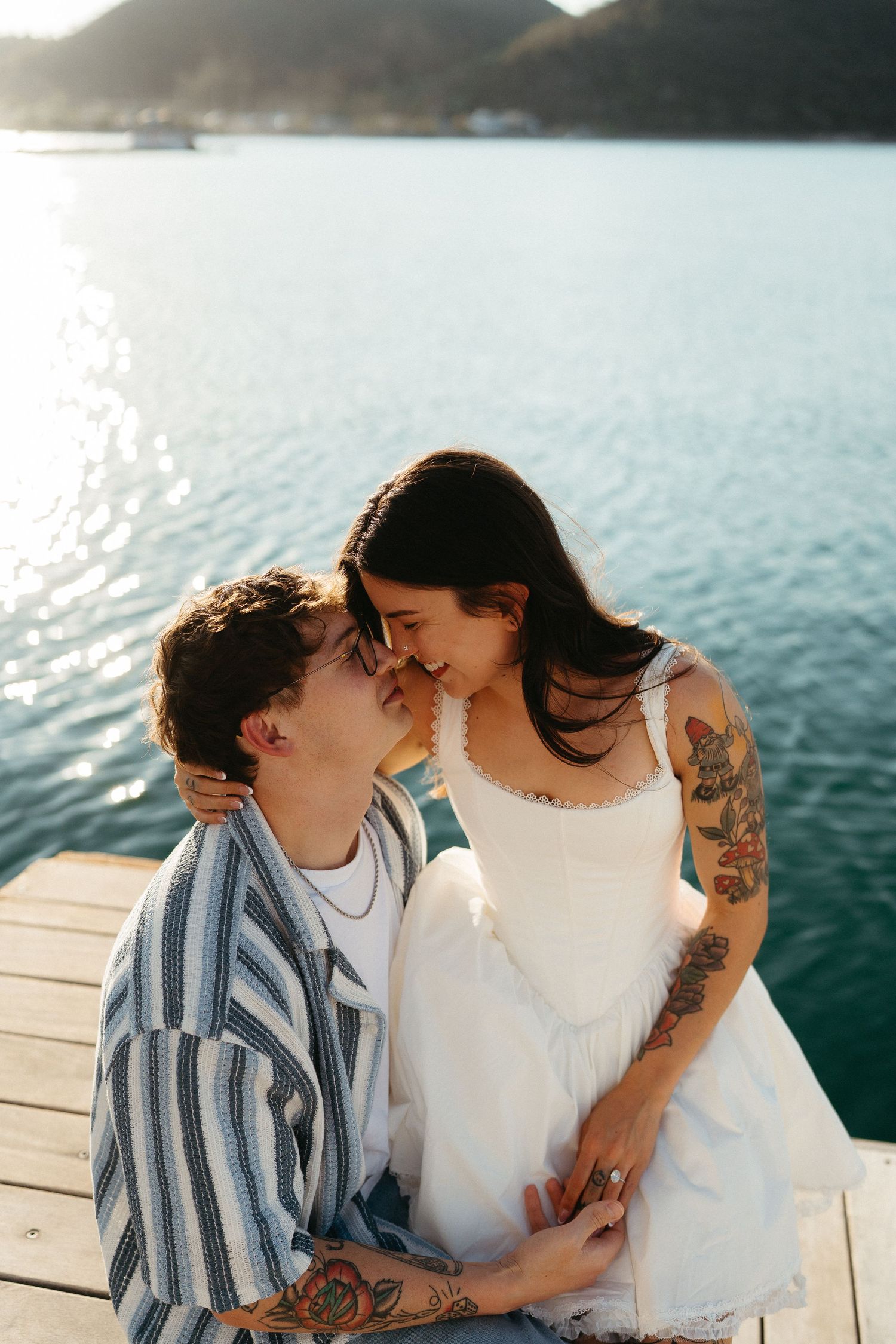A romantic couple sit close together on a wooden dock by a calm lake during sunset.