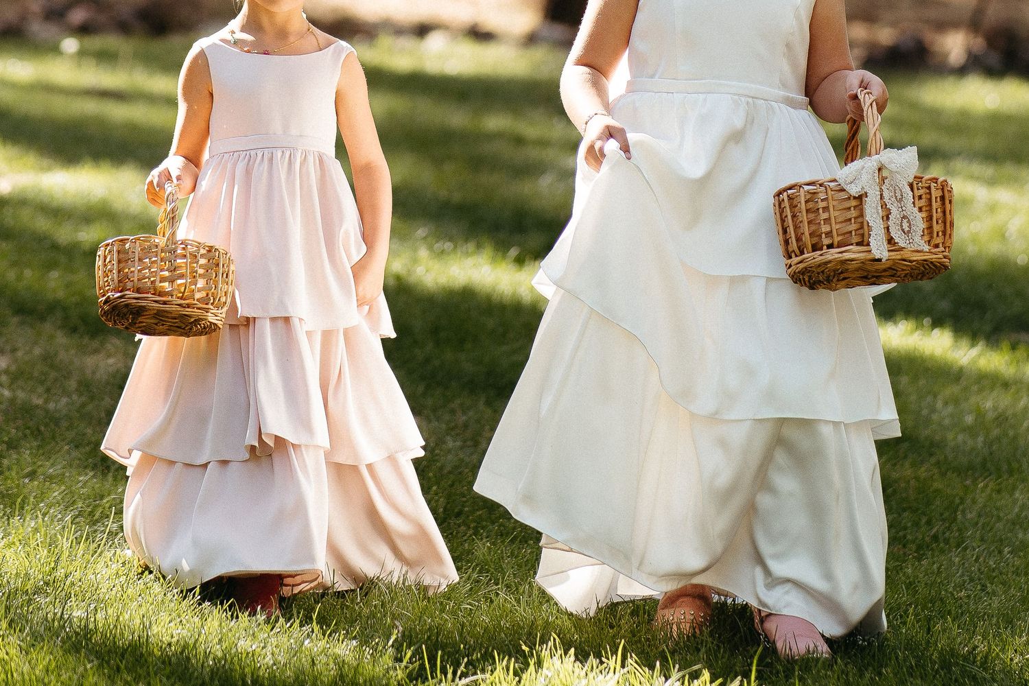 Two flower girls in white dresses walk on grass while carrying woven baskets at an outdoor wedding ceremony.