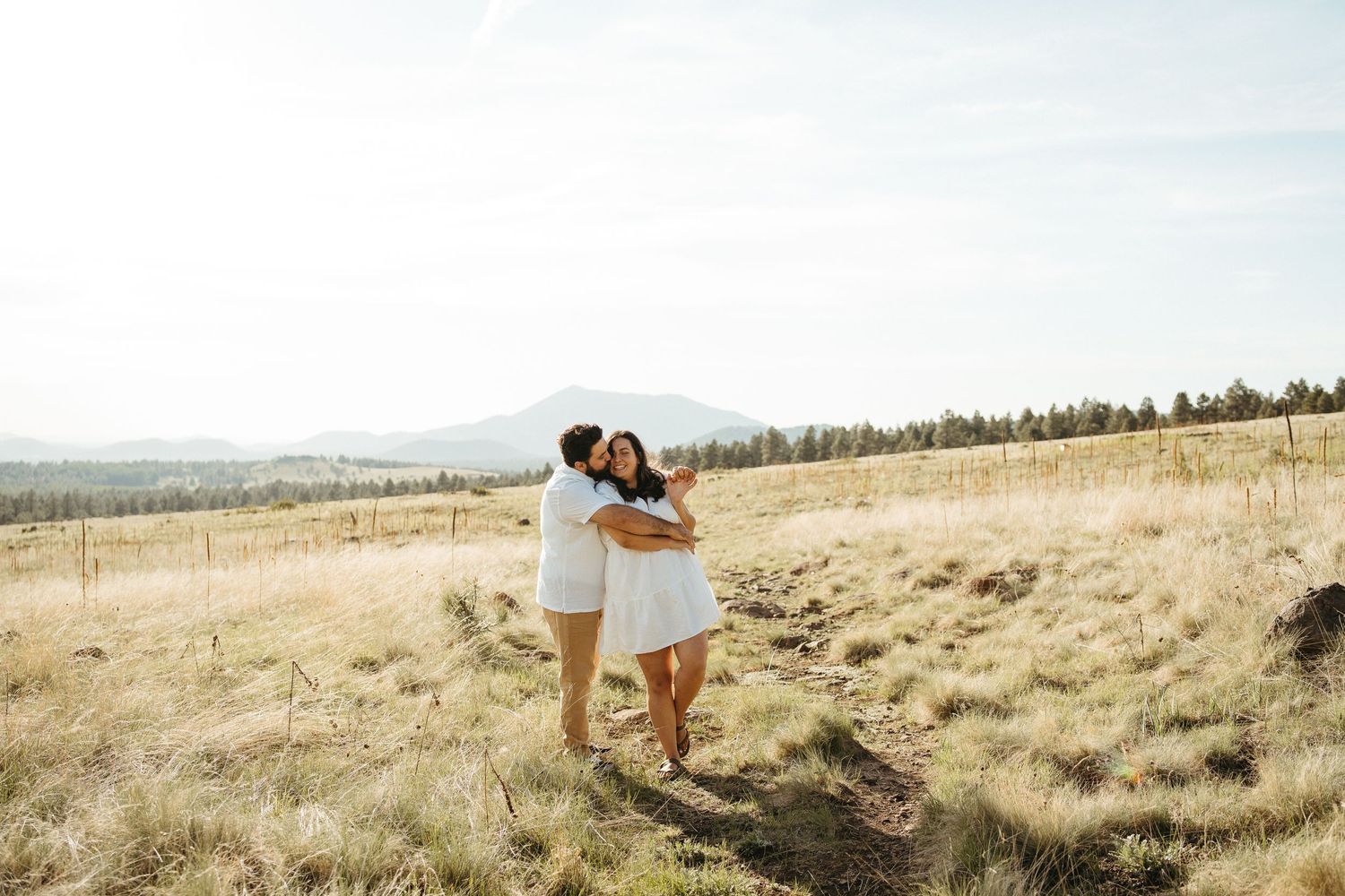 Couple in white outfits embracing on hillside with mountain vista in background.