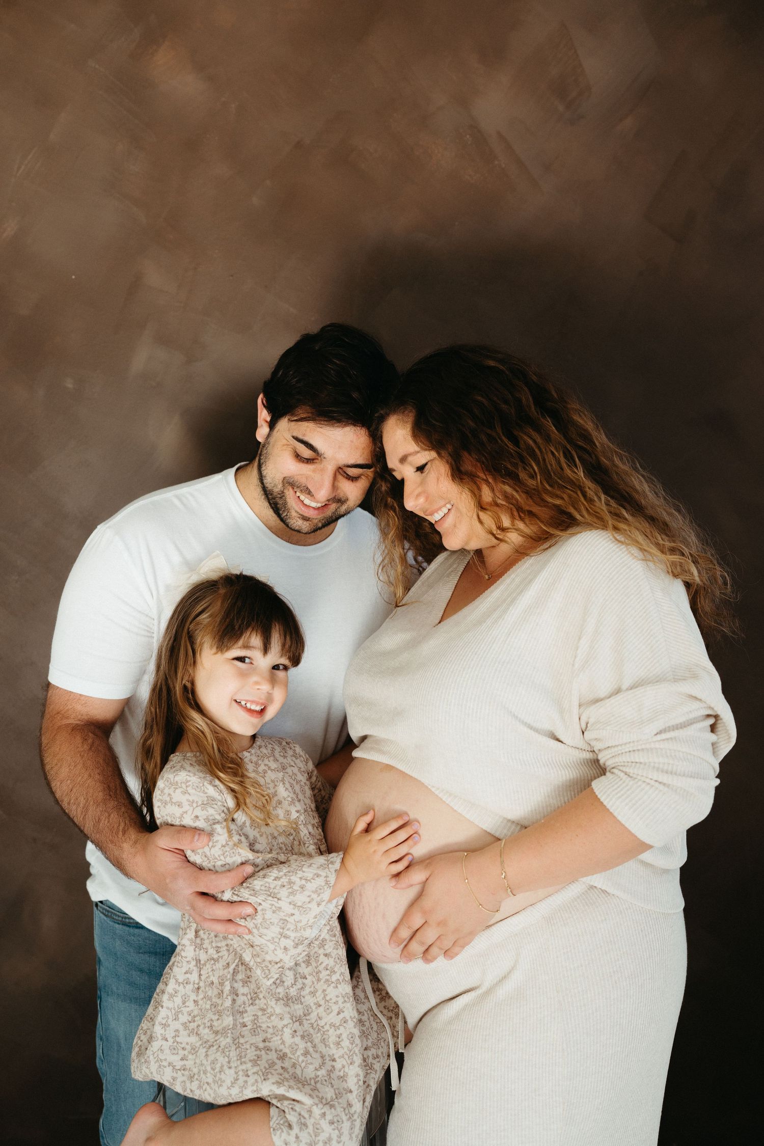 A pregnant woman in a white dress shares a tender moment with another person and small child against a dark background.