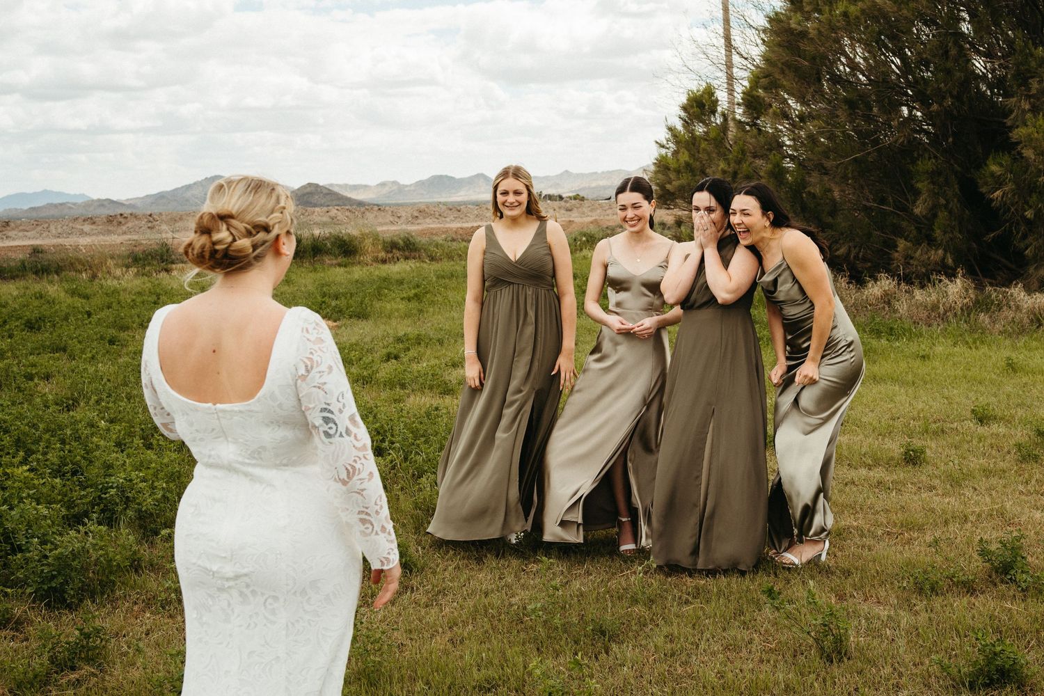 Bridesmaids in light brown dresses react joyfully on a grassy hill during an outdoor wedding ceremony.