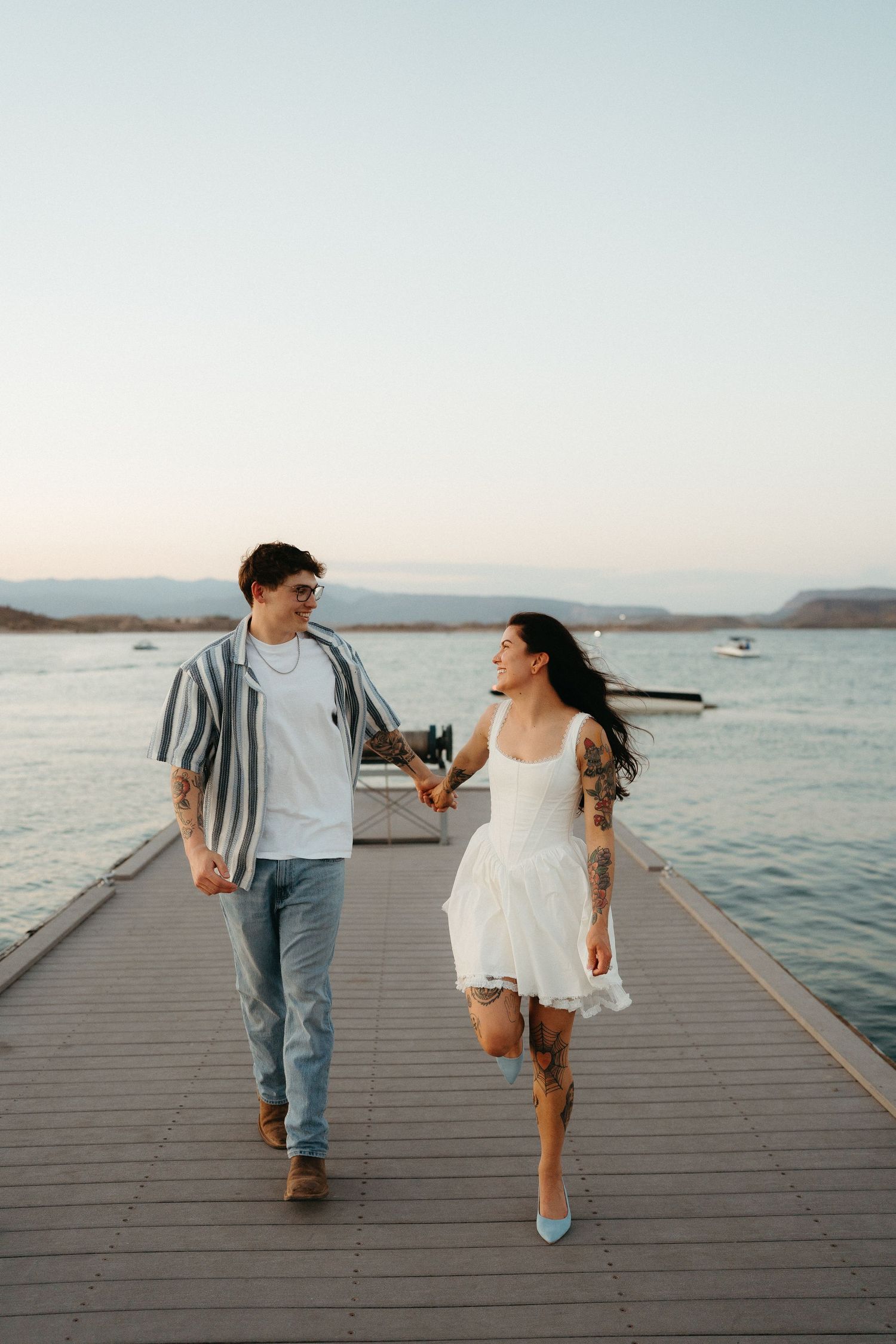 Two people walk hand in hand along a wooden dock by the water during sunset, creating a romantic silhouette.
