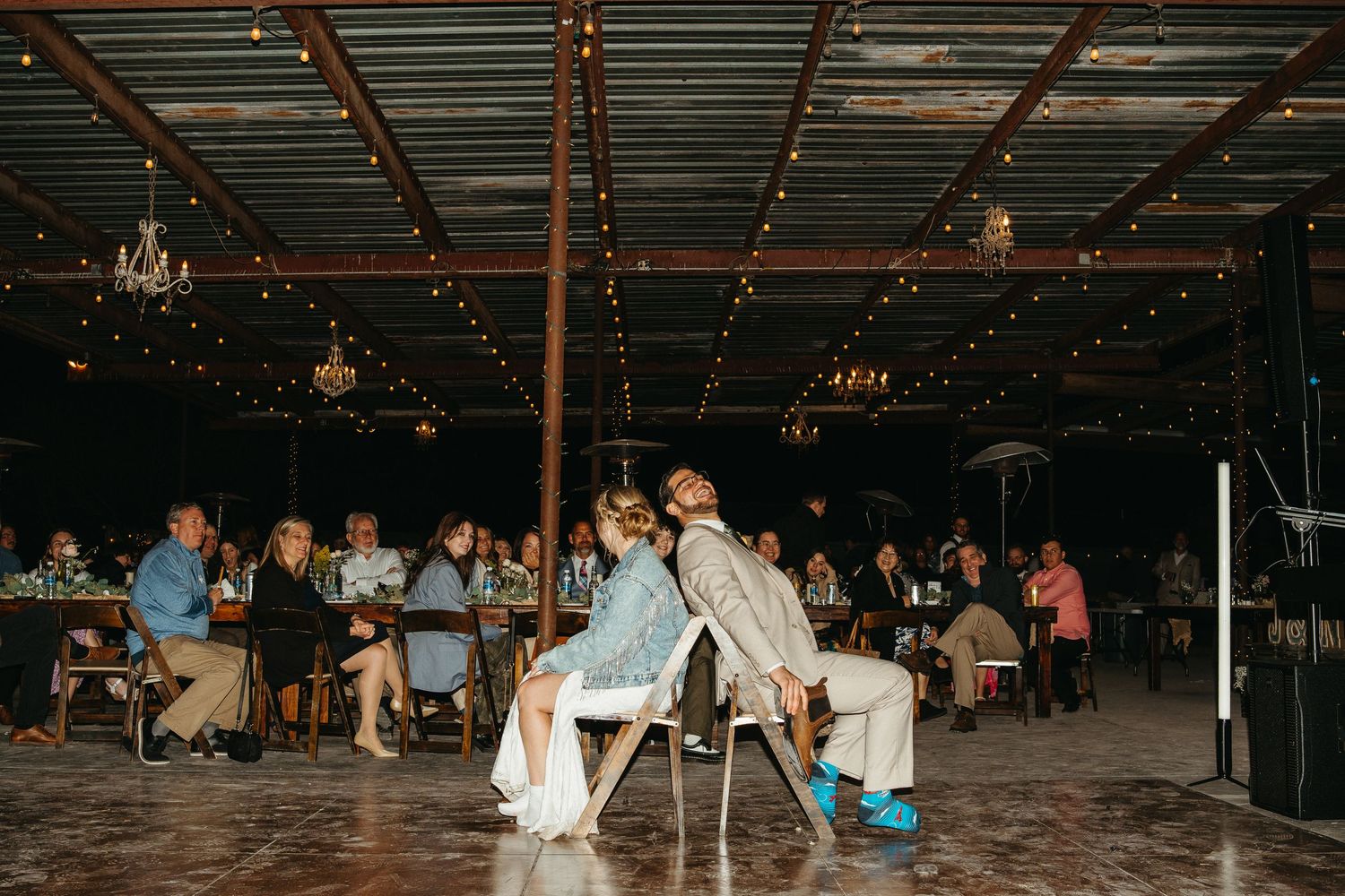 Wedding reception guests seated at tables in a rustic indoor venue with string lights overhead.
