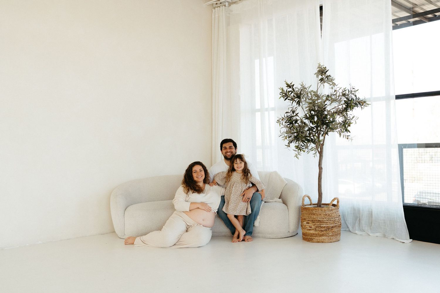 A small family shares an intimate moment together on a white curved sofa in a bright minimalist room with sheer curtains.