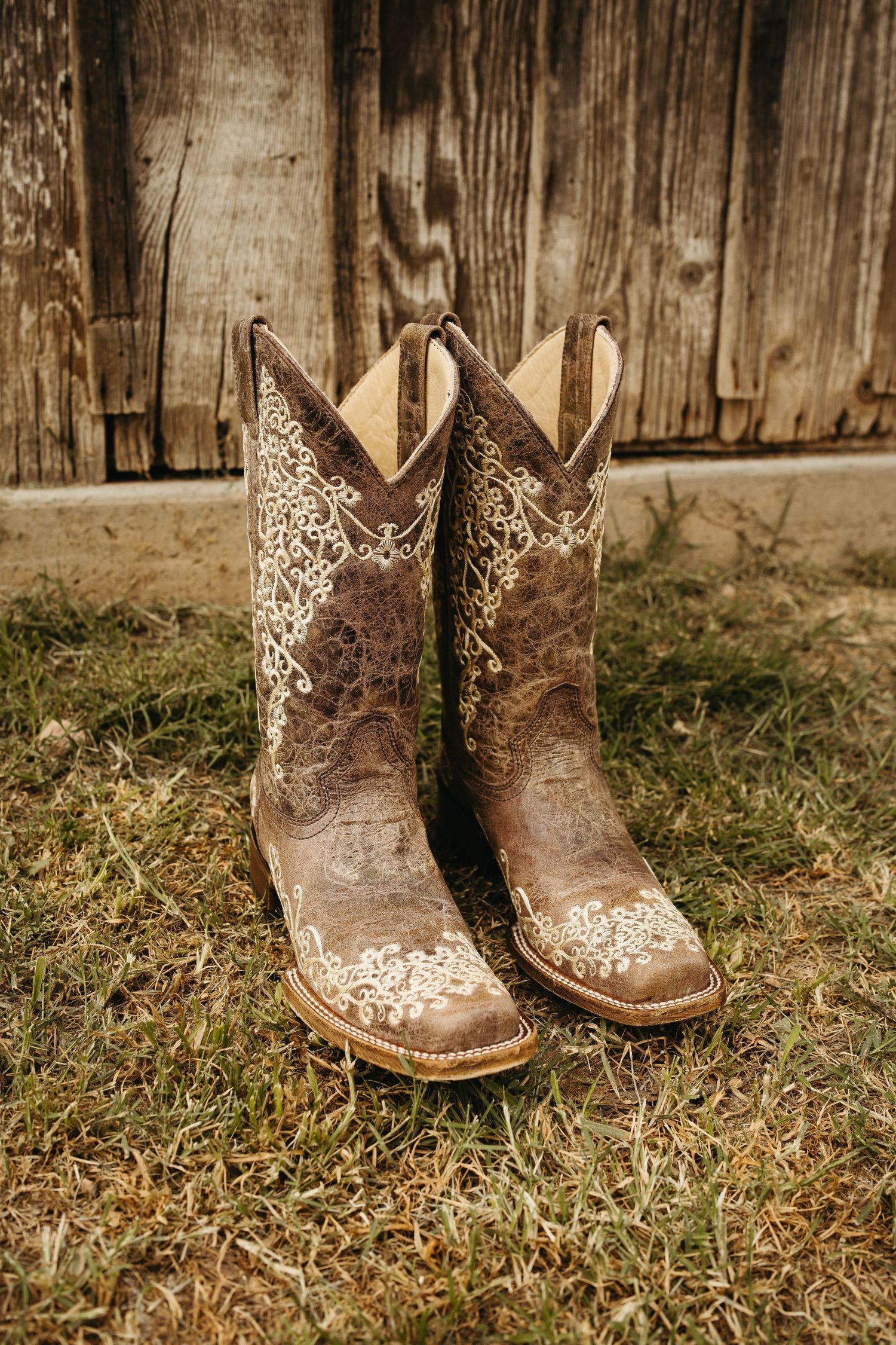 Brown leather cowboy boots with intricate cream floral embroidery displayed on grass.