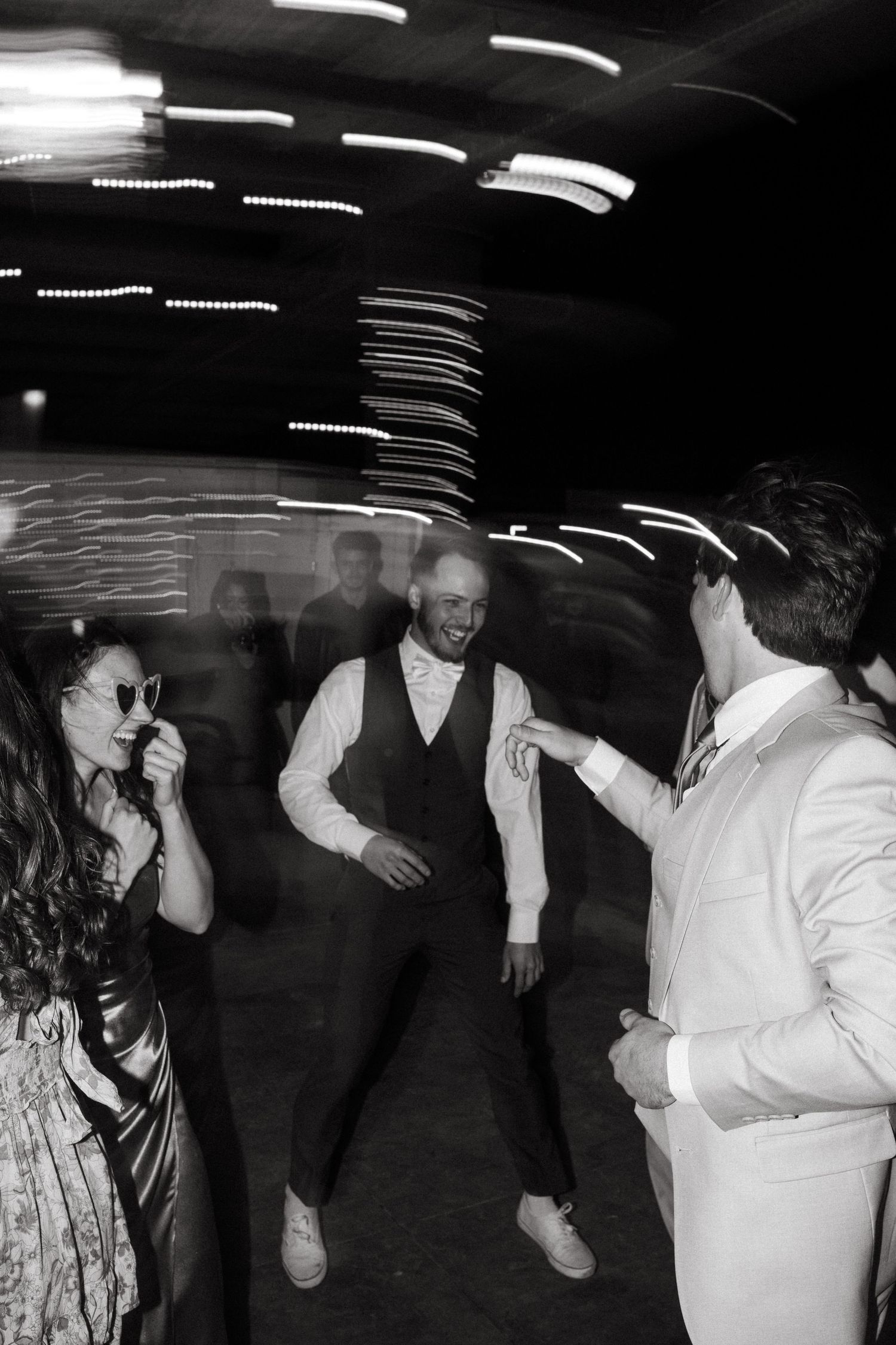 A black and white photo of people dancing at a reception under geometric ceiling lights.