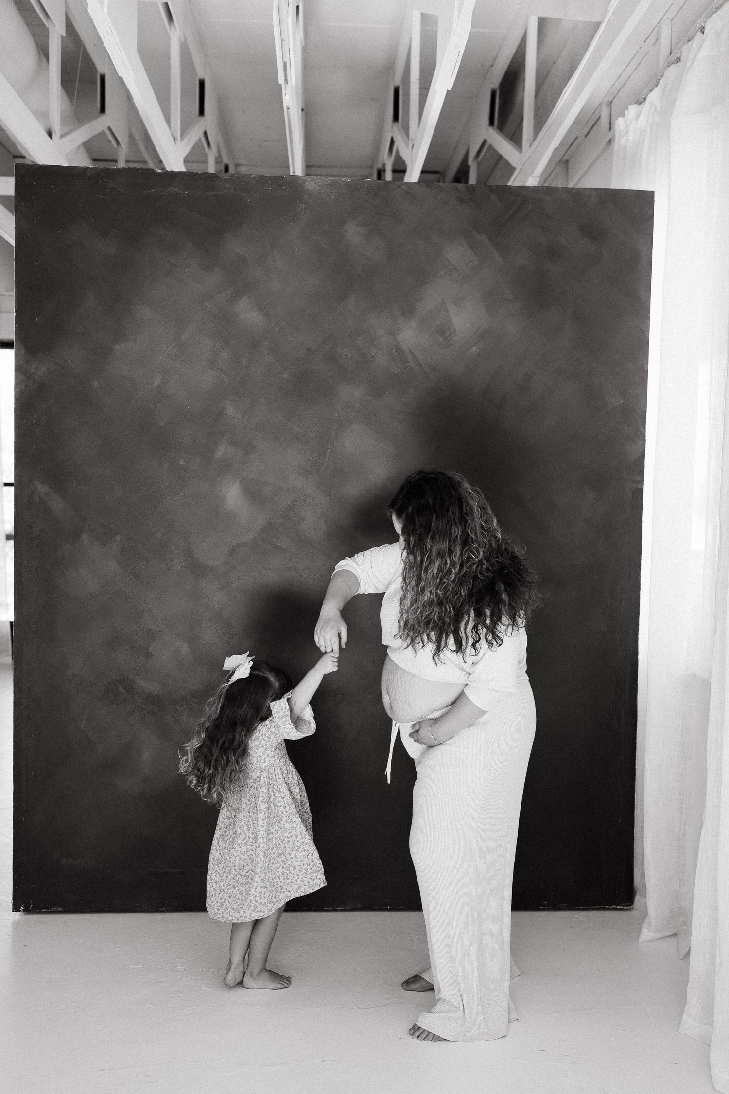A black and white photo of a parent and child dancing together in front of a dark backdrop in a bright studio space.
