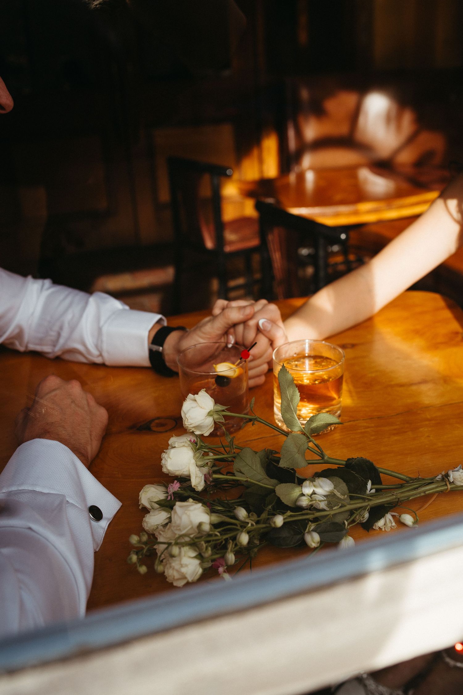 White roses and drinks on wooden table with hands clasped in warm lighting.