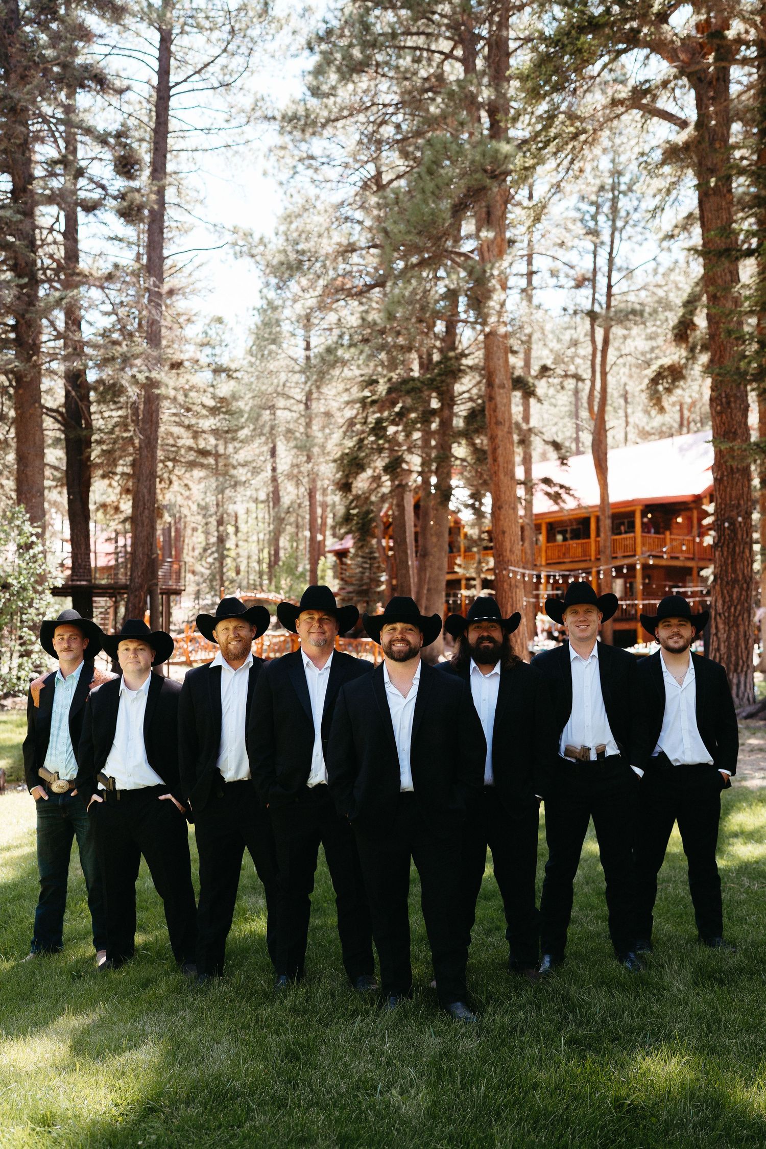 A group in matching black suits and cowboy hats stand in formation on grass with tall pine trees and a cabin in background.
