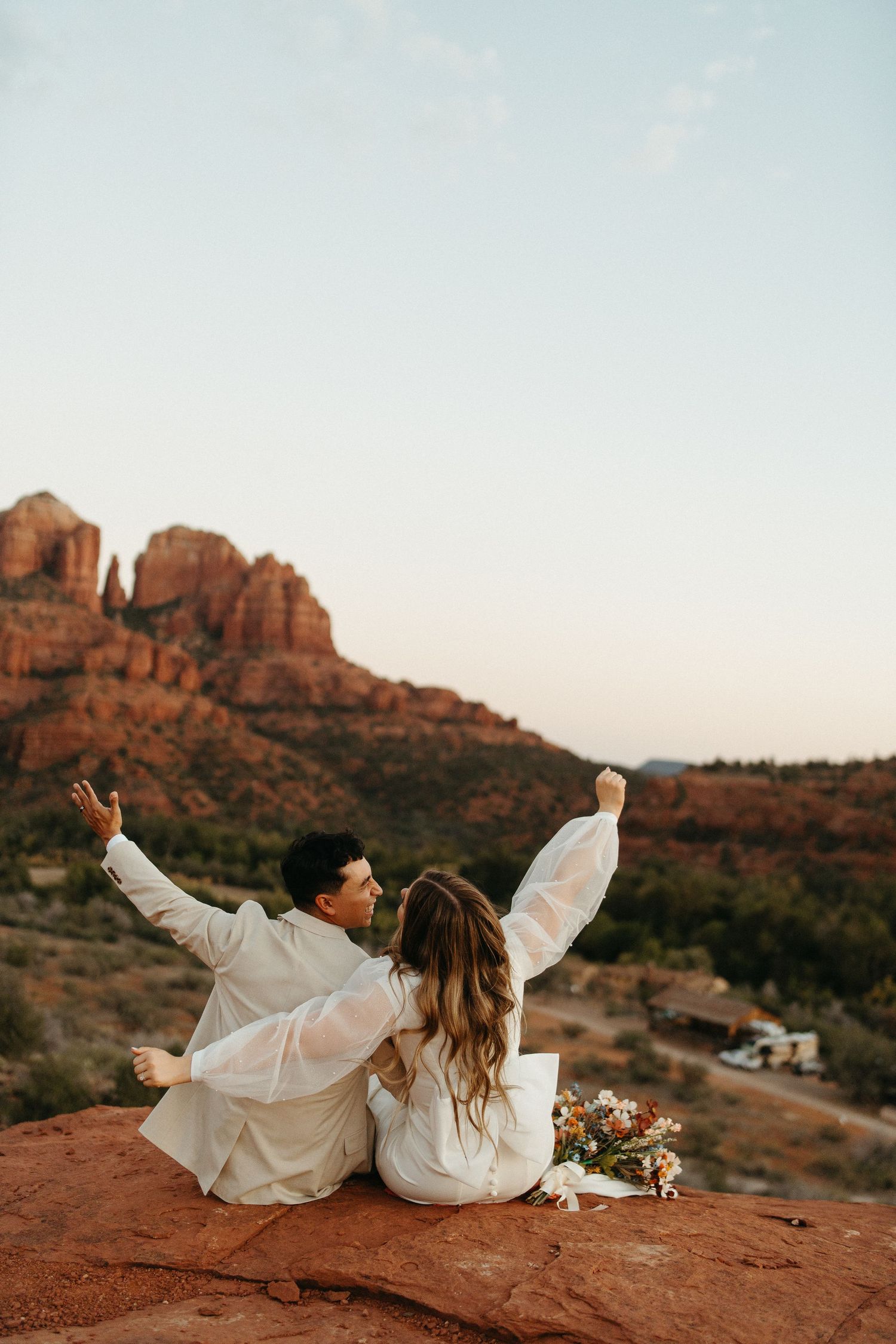 Couple in white outfits sitting on red rocks with scenic desert landscape backdrop.