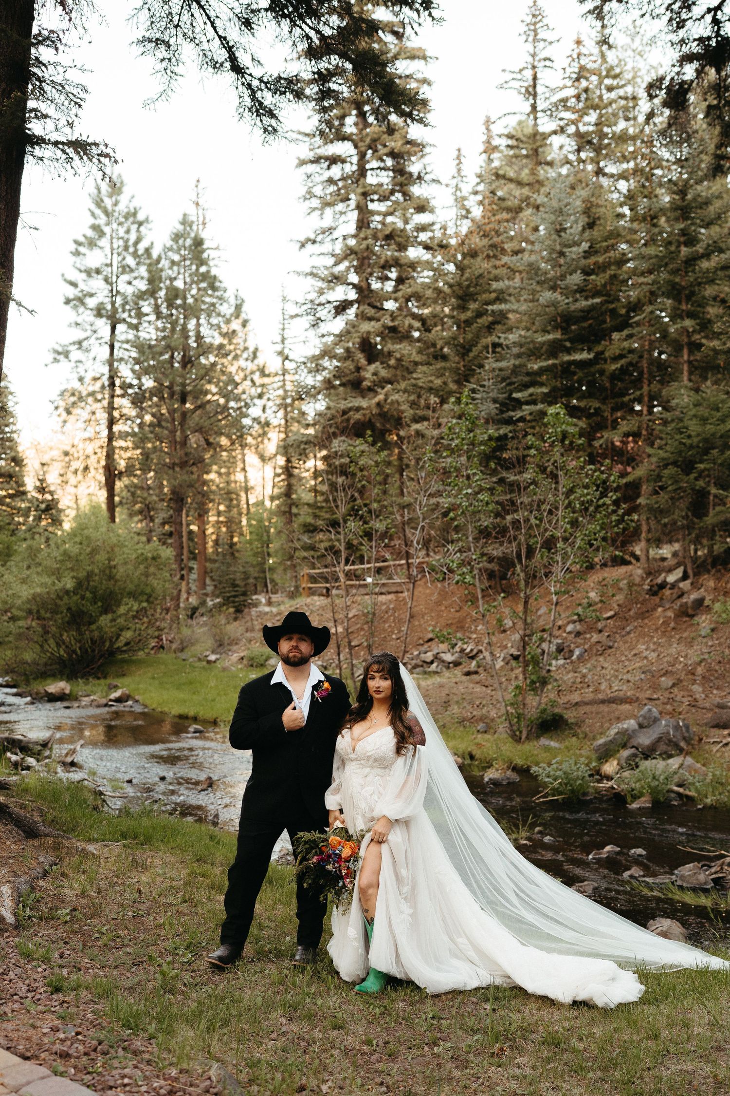 A couple in wedding attire poses together by a scenic forest stream during their outdoor ceremony.