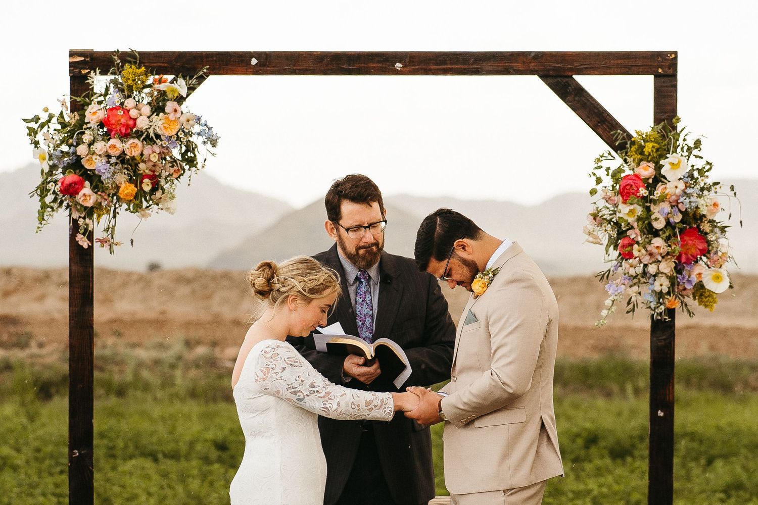 Intimate outdoor wedding ceremony beneath rustic wooden arch decorated with colorful flowers.