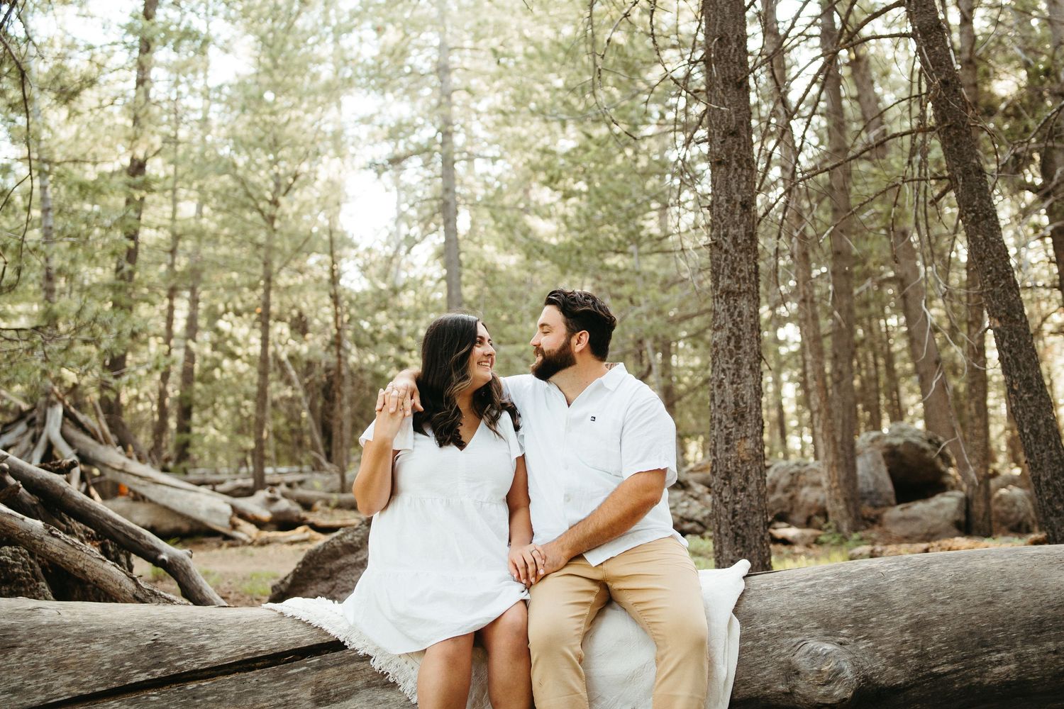 Couple in coordinated white outfits sitting on fallen log in forest setting.
