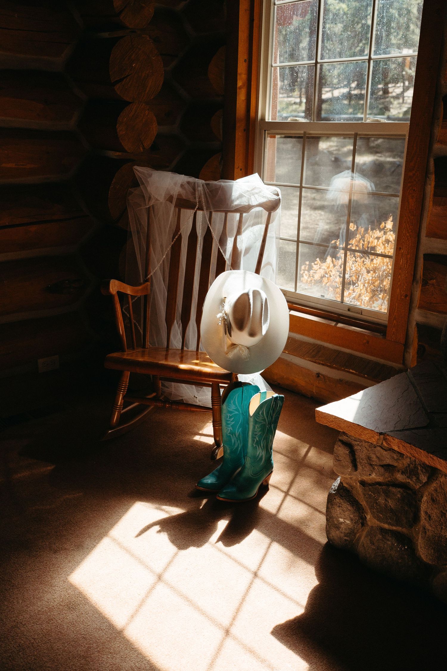 Sunlight streams through cabin window onto wooden rocking chair with cowboy hat and turquoise boots in rustic interior.
