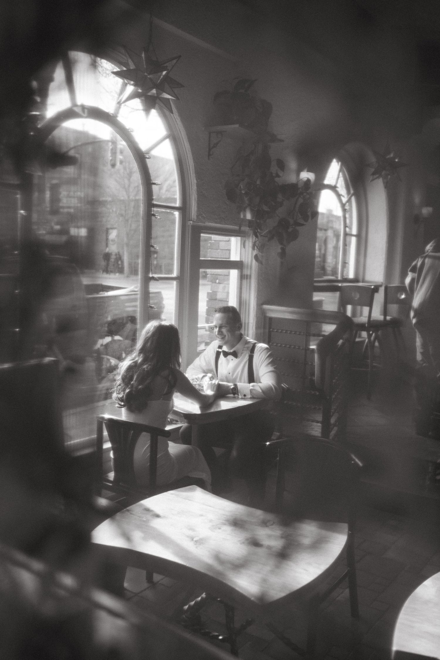 Black and white photo of couple at cafe table beneath arched windows.