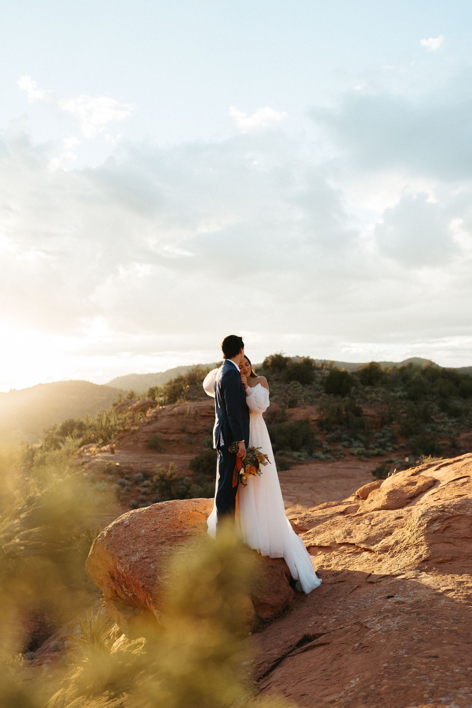 A couple in wedding attire embraces on a red rock cliff at sunset with dramatic desert landscape in the background.