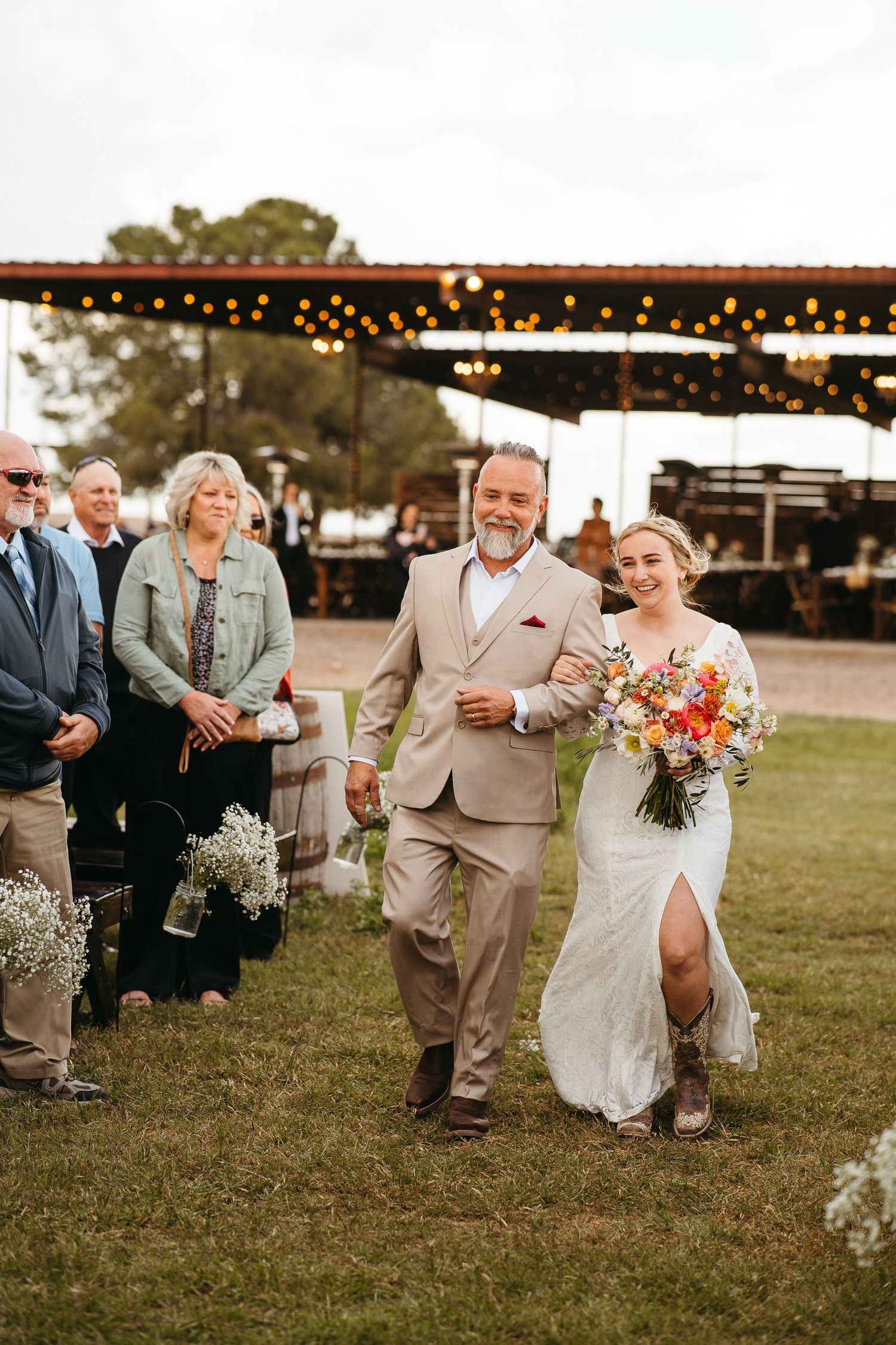 Wedding couple walks down aisle under string lights during outdoor ceremony.