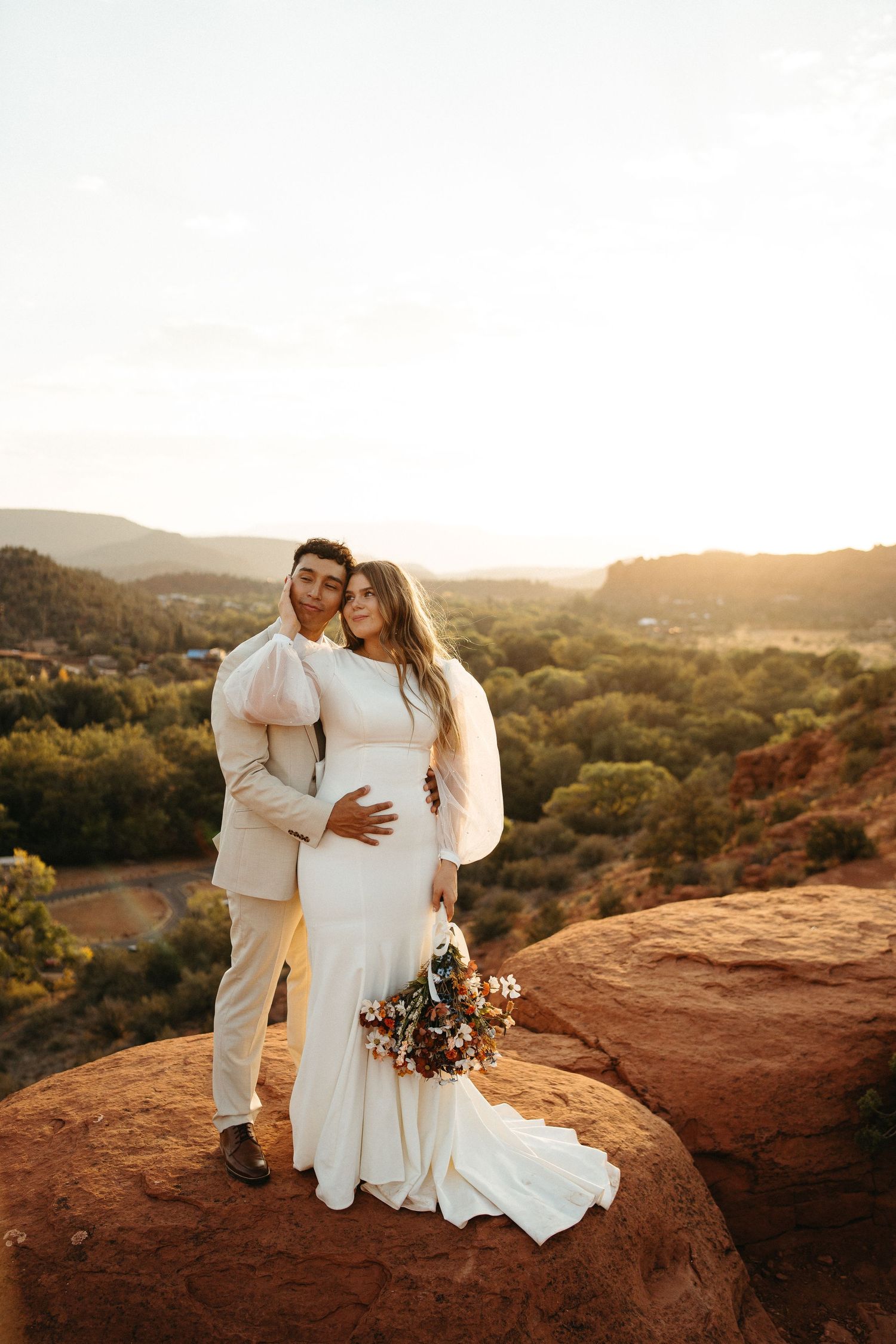 Wedding couple embracing on red rocks overlooking scenic mountain landscape at sunset.