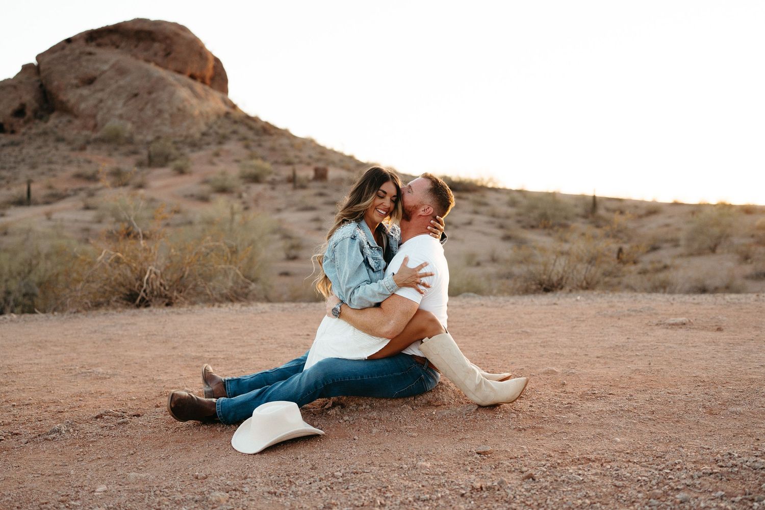 A couple in western wear sits intimately on desert ground with rocky terrain behind them.