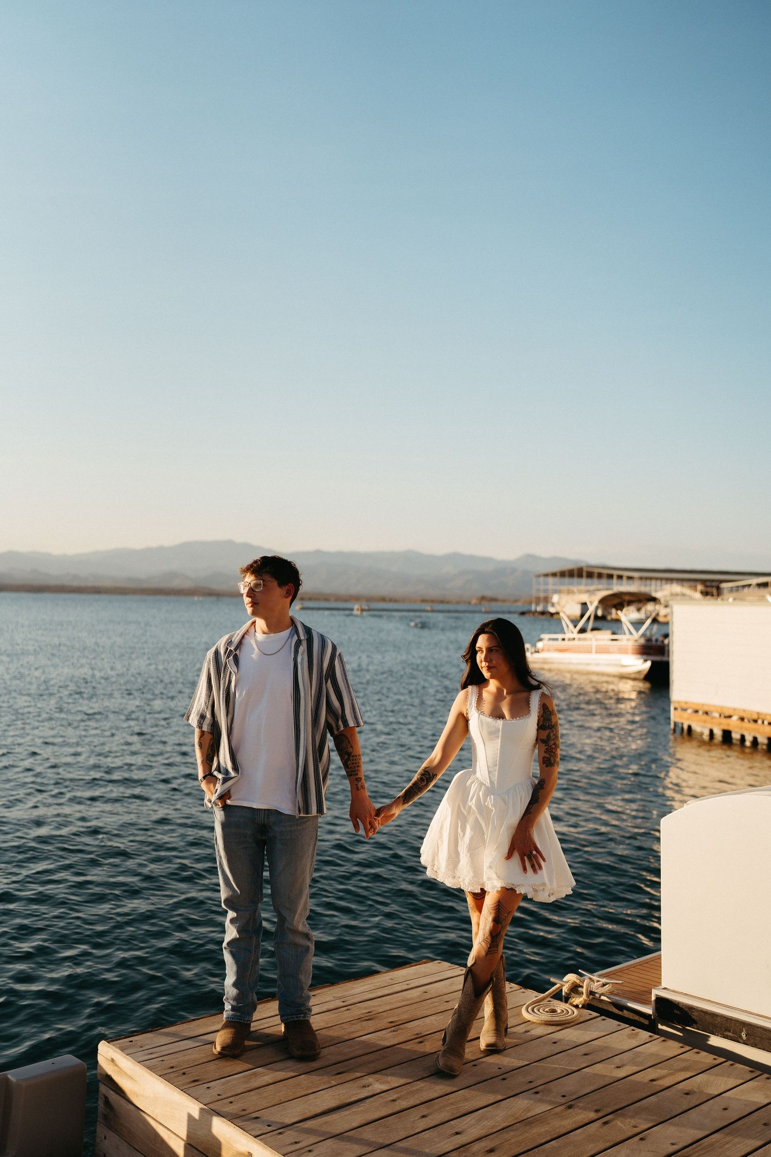 A couple holds hands while walking on a wooden dock by a lake with mountains in the background at sunset.