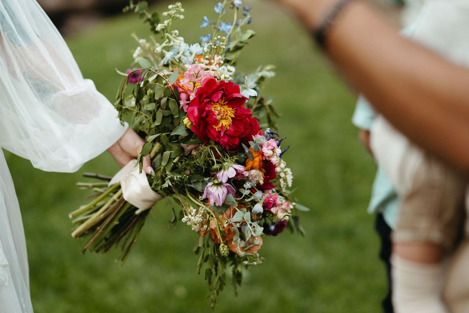 A rustic wildflower wedding bouquet with red, purple and white blooms held against a green grass background.