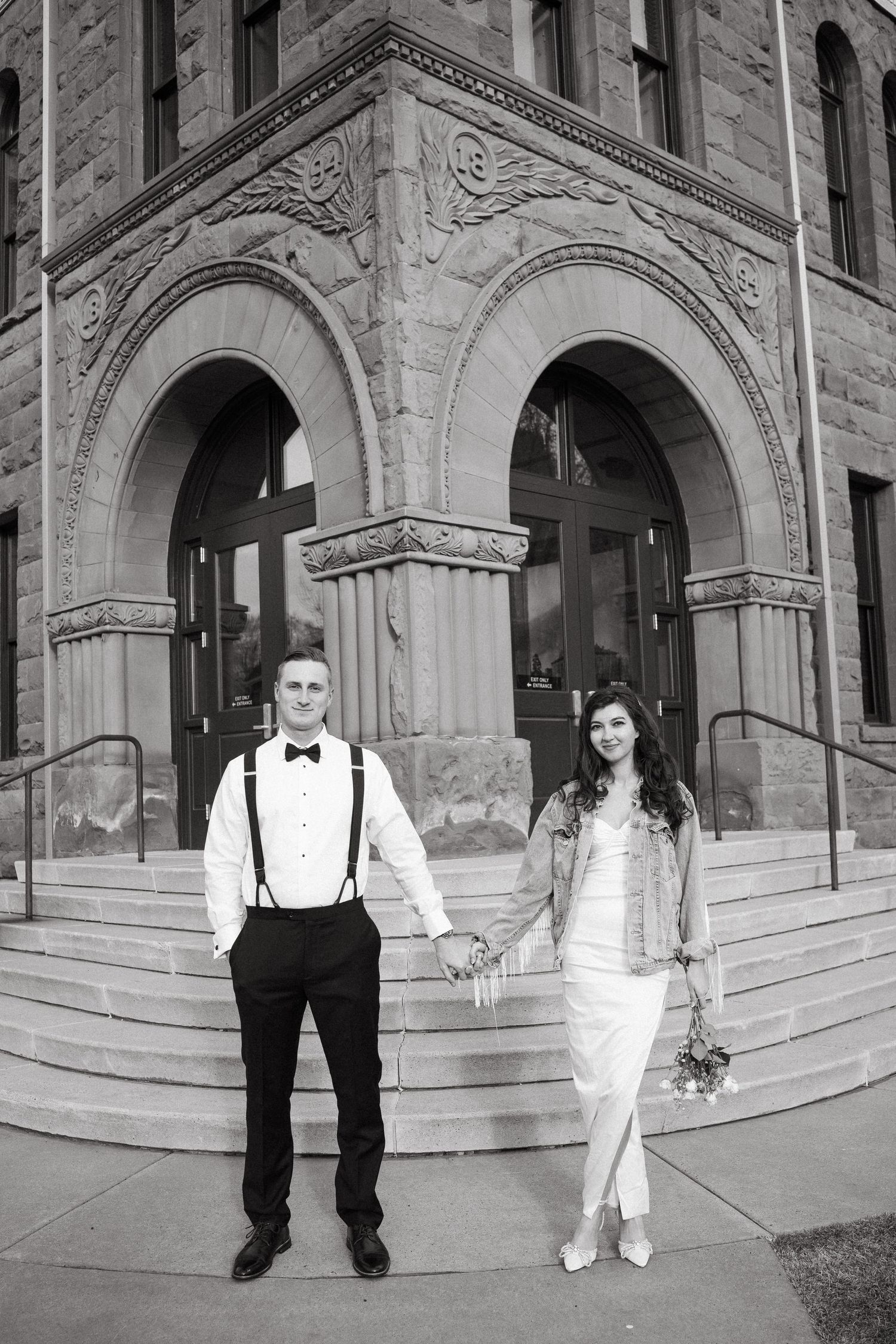 A formal couple poses in front of an ornate stone building with arched windows.