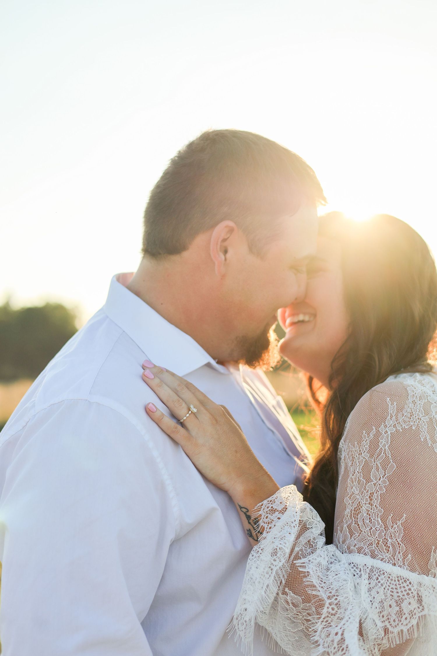 A romantic couple embraces in golden sunlight, with the bride wearing lace sleeves and a sparkling engagement ring.