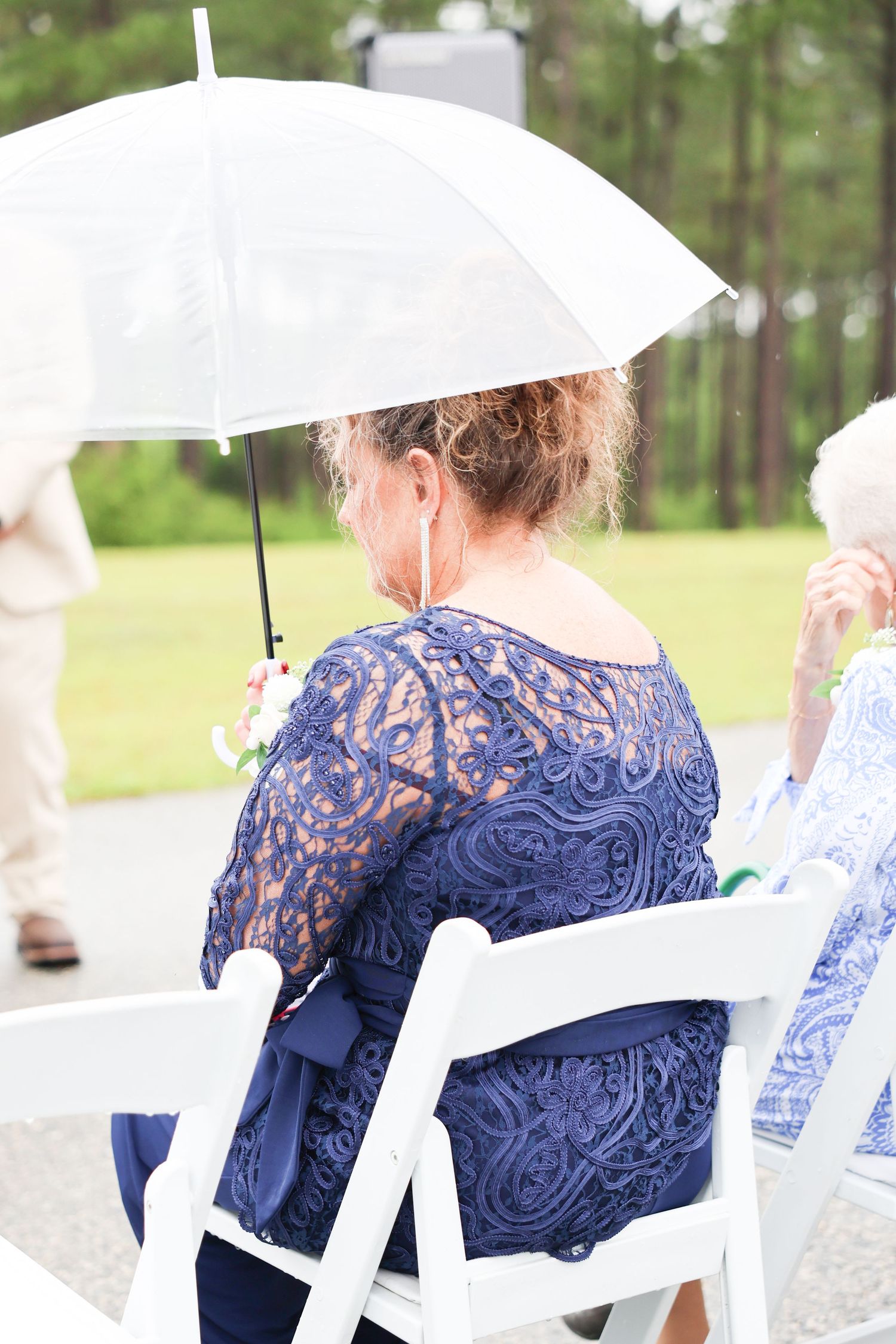 A wedding guest in a navy lace dress holds a clear umbrella while seated at an outdoor rainy ceremony.
