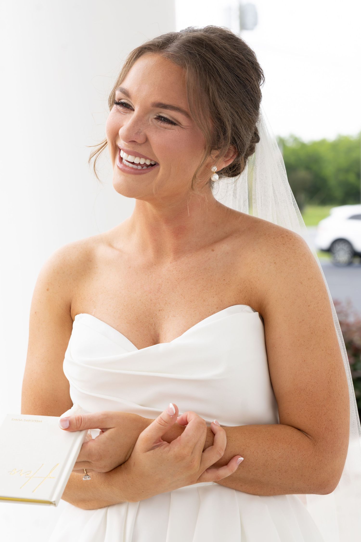 Smiling bride in strapless white gown holding a book, wearing a veil and pearl earrings on her wedding day.