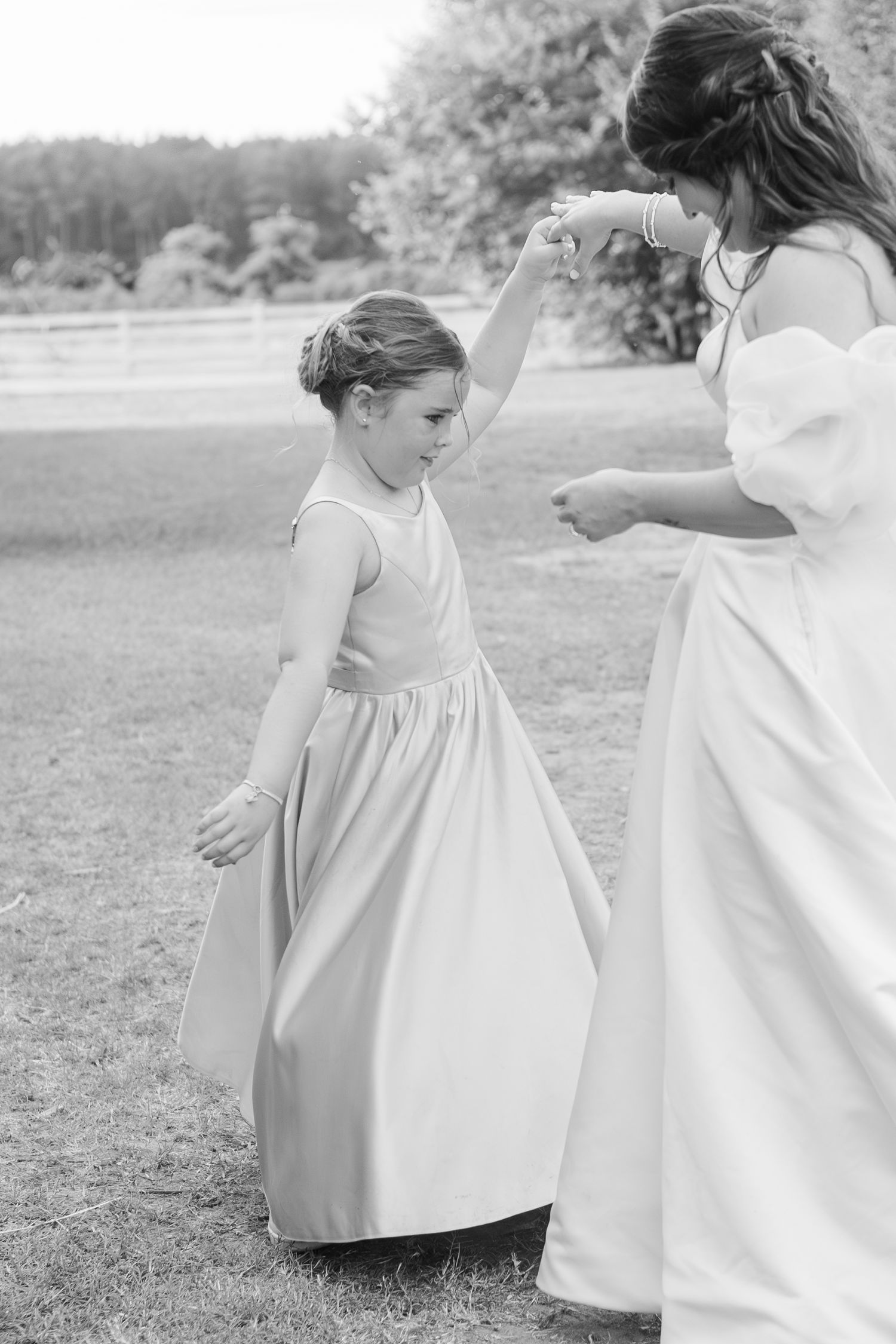 Bride twirling flower girl in long satin dress on green lawn at outdoor wedding ceremony in black and white photo.