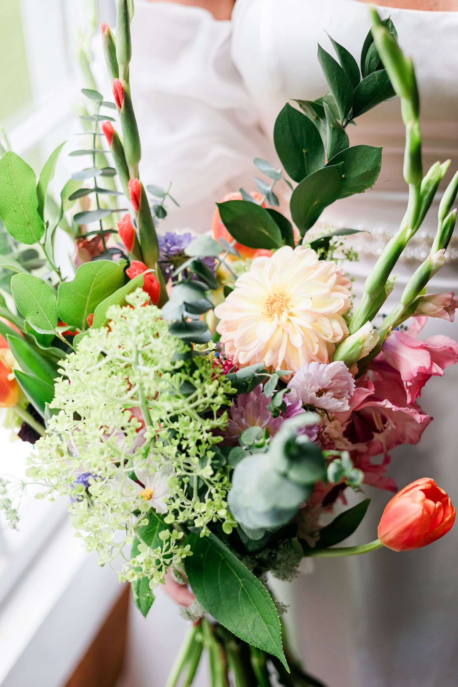 Colorful wedding bouquet featuring peach dahlia, red tulips, pink flowers, and lush greenery held by a bride.