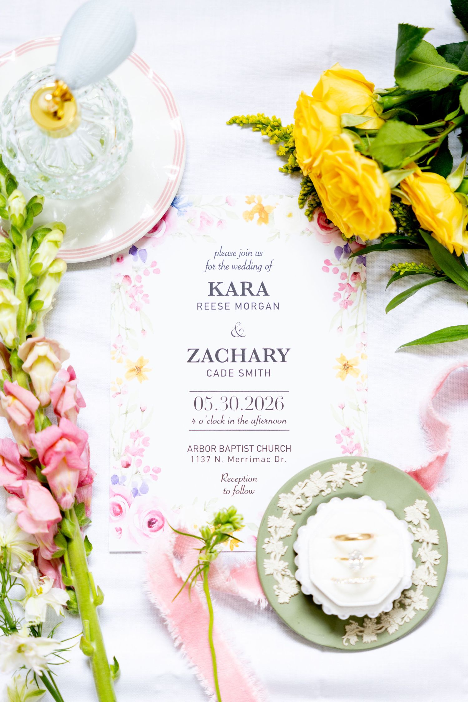 Elegant wedding invitation for Kara and Zachary surrounded by yellow roses, pink flowers, perfume bottle, and gold rings.