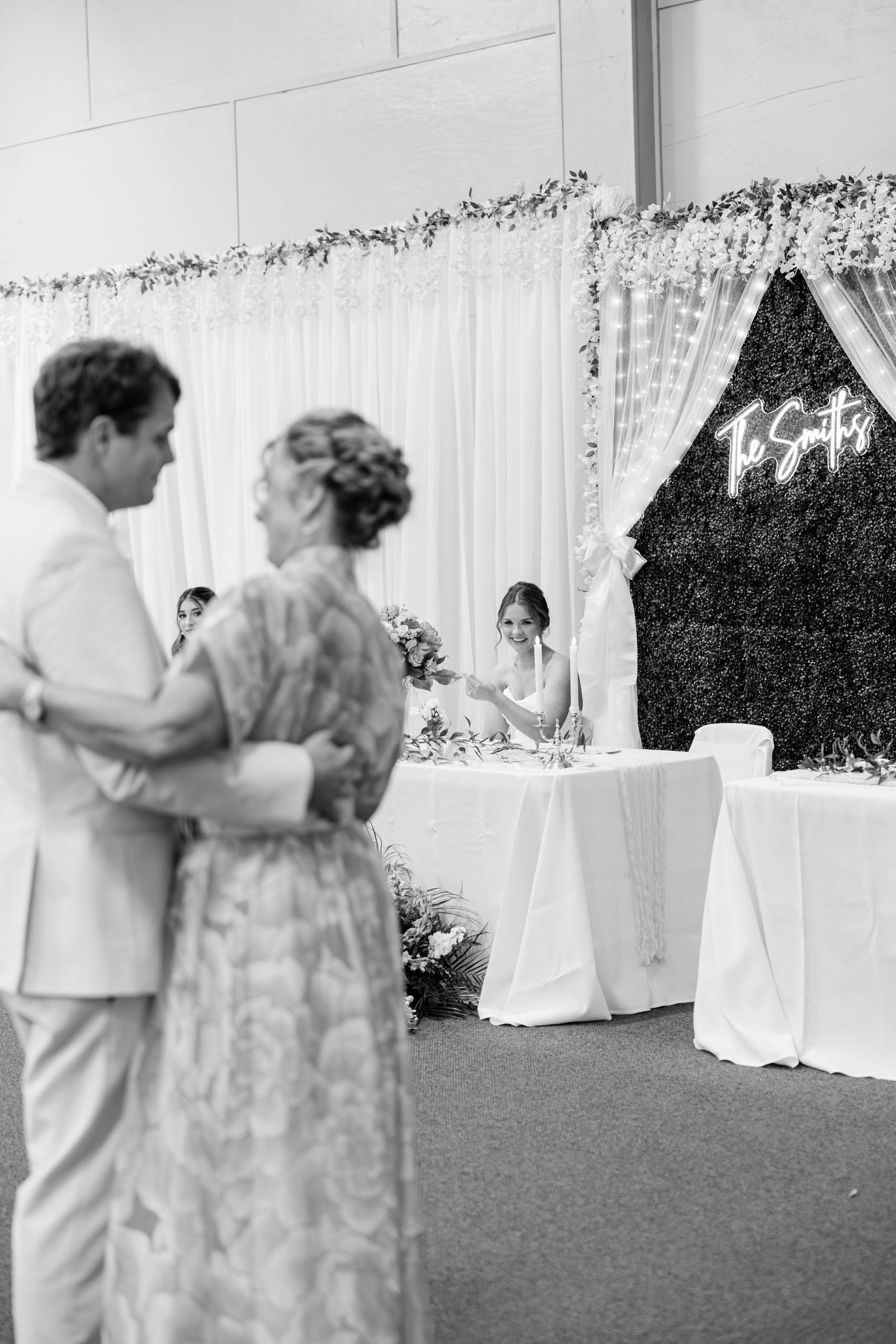 Black and white photo of couple watching bride at wedding reception with floral backdrop and neon Smith sign.