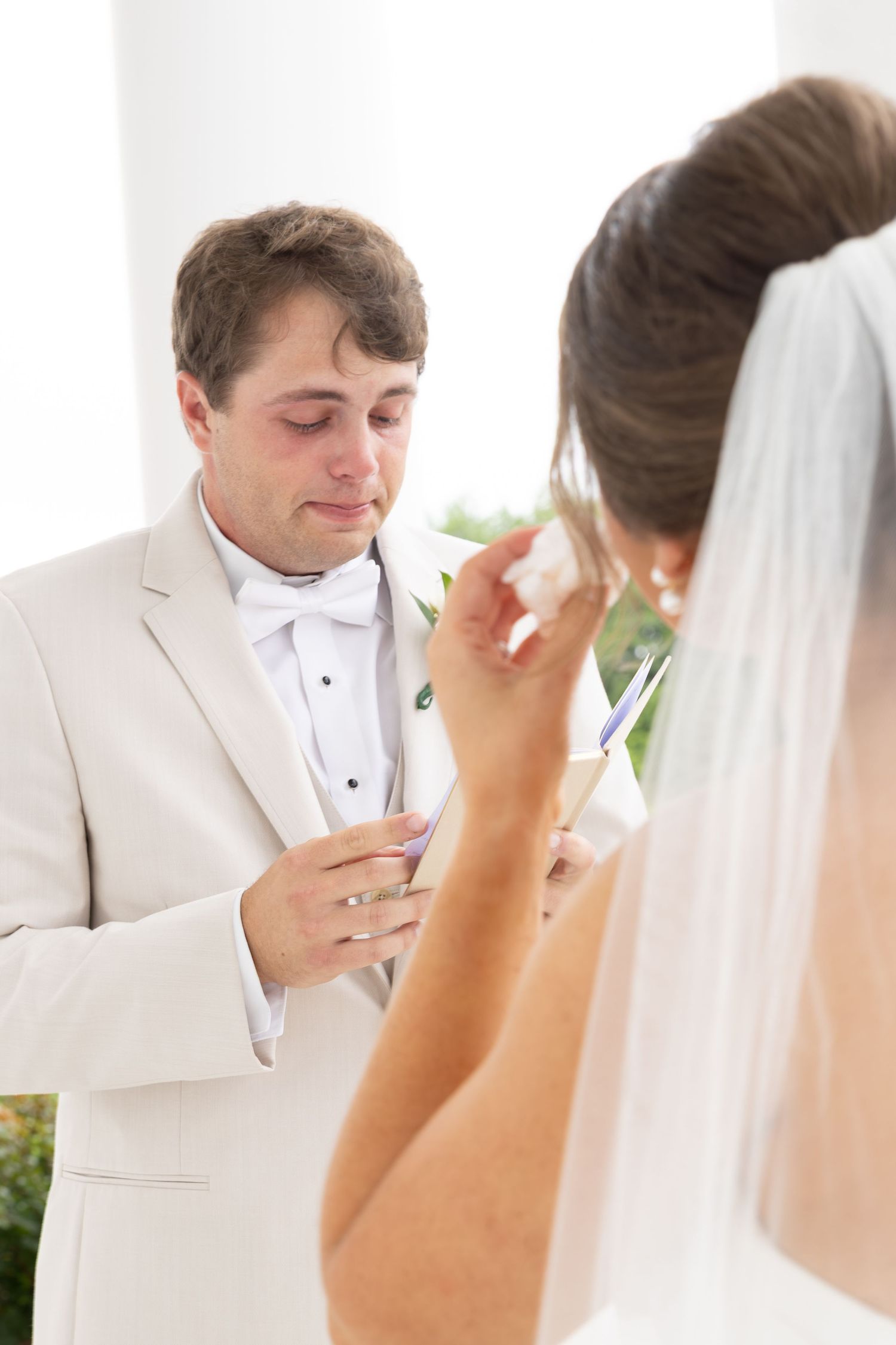 Tearful groom in cream tuxedo reading wedding vows while bride in veil stands before him.